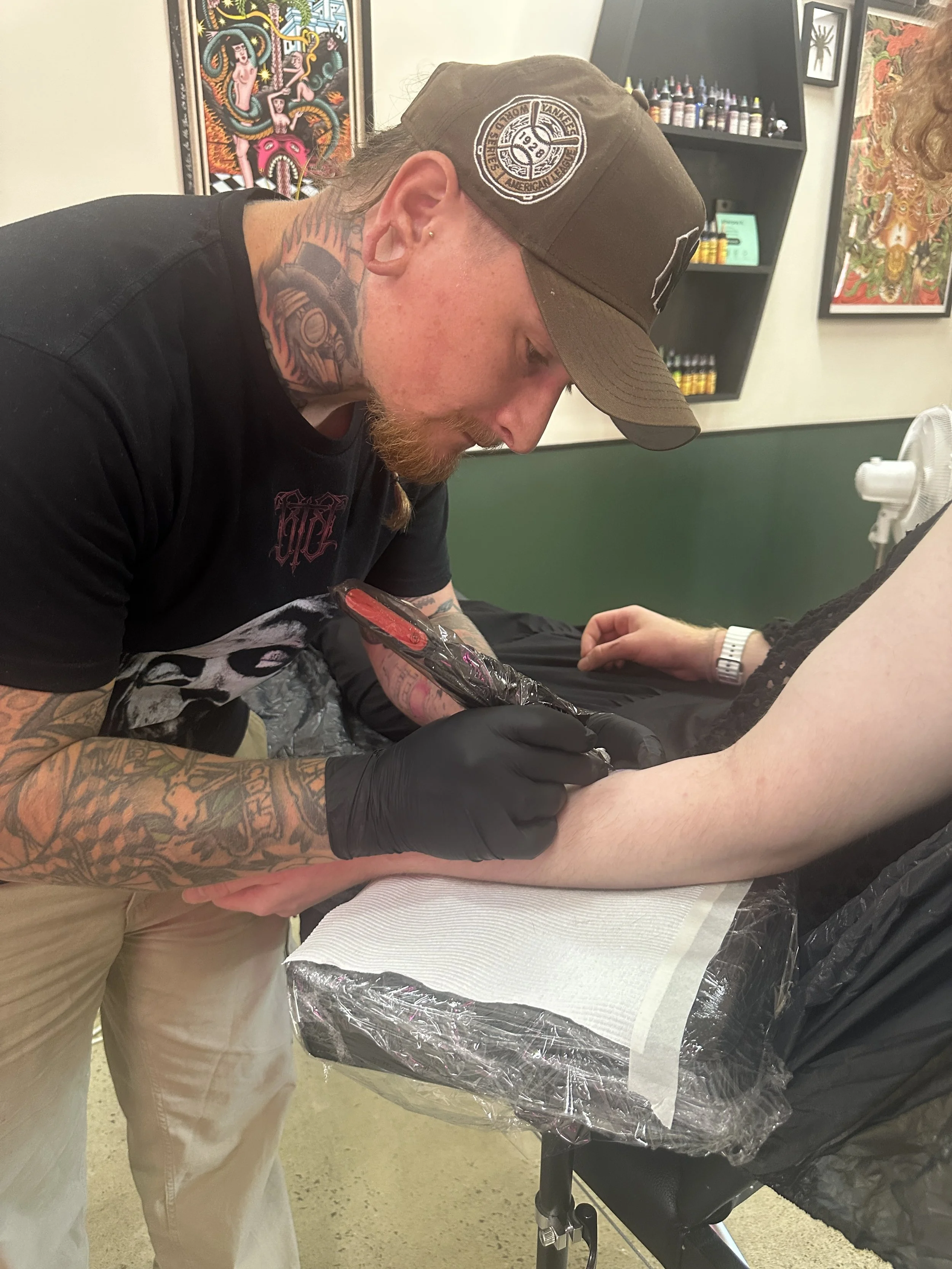 A man with tattoos on his right arm and neck, wearing a black T-shirt and a brown cap, is getting a tattoo on his left forearm. Another person is assisting, wearing a white watch. The background features artwork on the walls, including a colorful painting and tattoo-related supplies on shelves.
