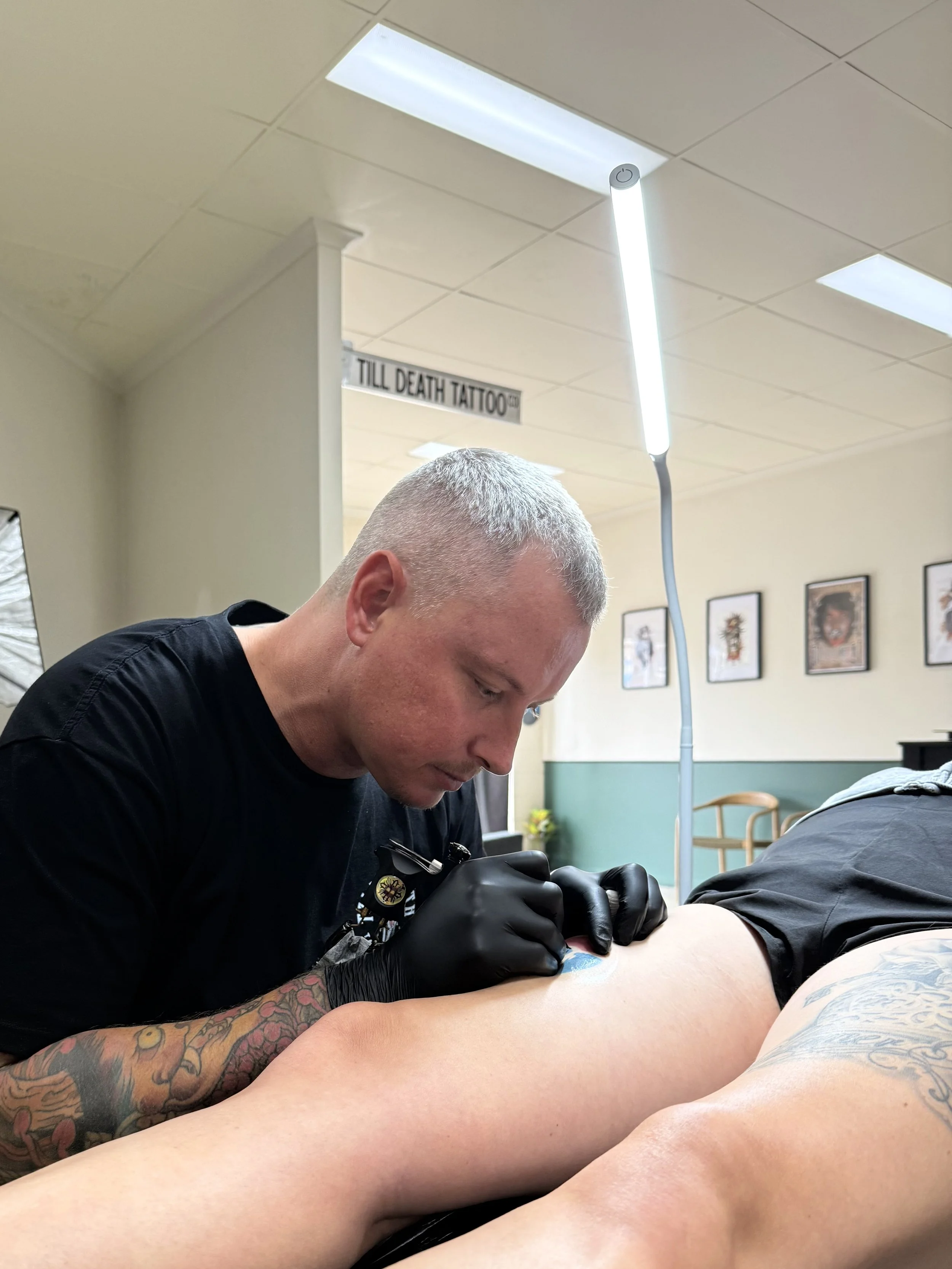 A tattoo artist is working on a client's arm in a studio with framed pictures on the wall. The artist wears black gloves and a black shirt, and there is a sign in the background that reads 'TILL DEATH TATTOO'.