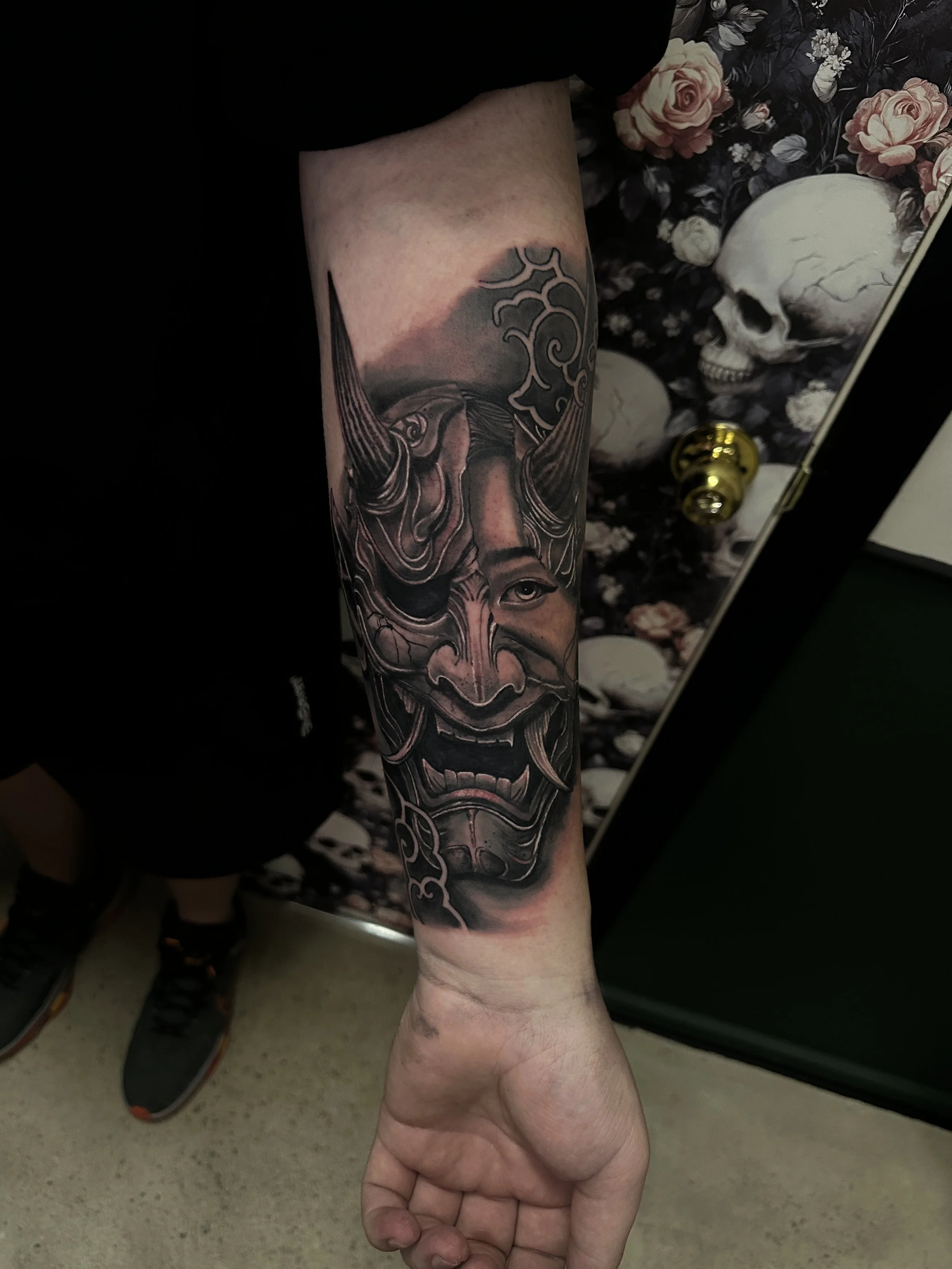 Tattoo of a detailed traditional Japanese mask on a person's forearm, with a floral background and a skull illustration partially visible behind.