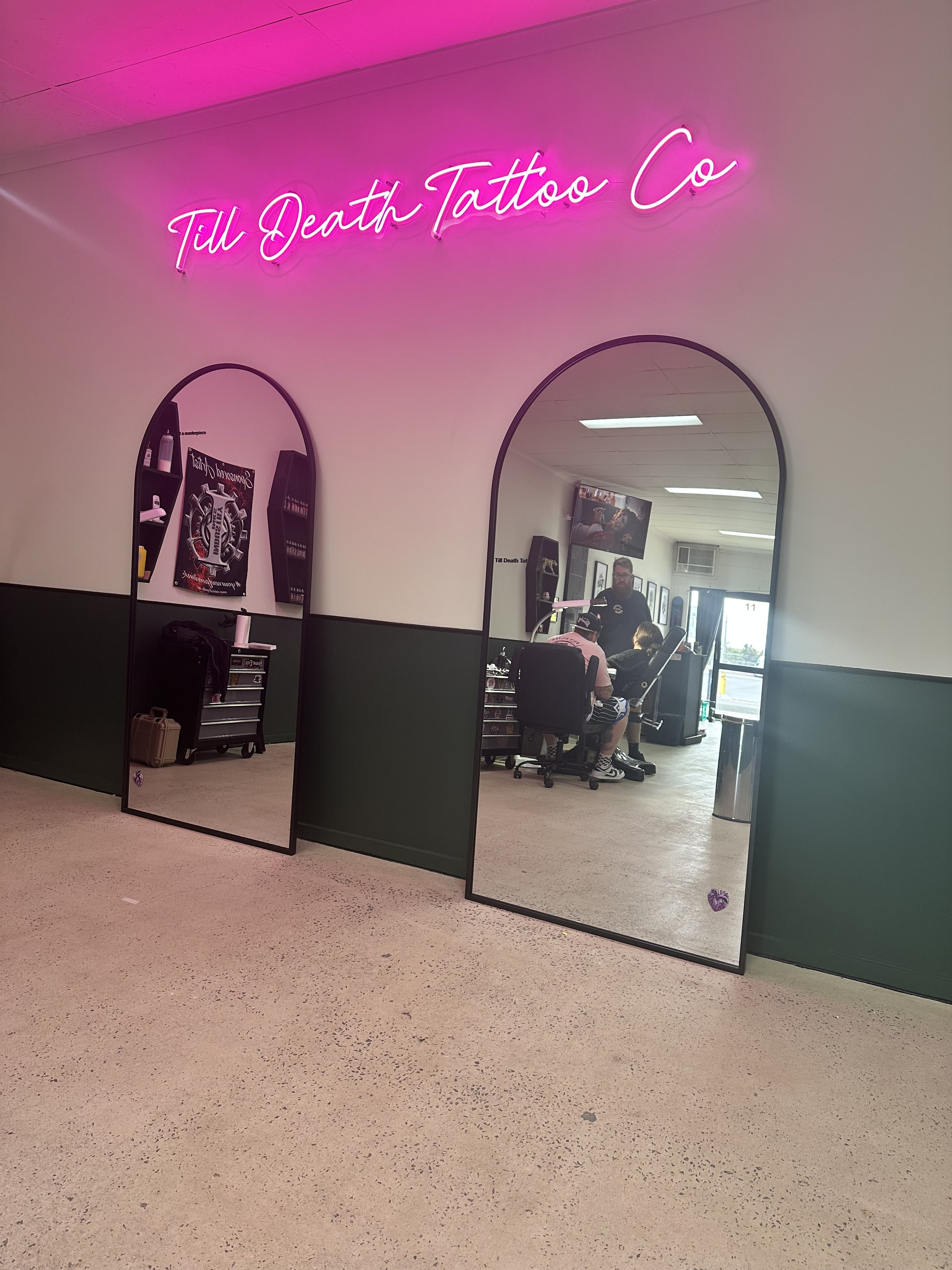 Inside a tattoo parlor with a pink neon sign that reads 'Til Death Tattoo Co'. There are two mirrors, one showing a tattoo artist working with a client, a television on the wall, and various tattoo supplies.