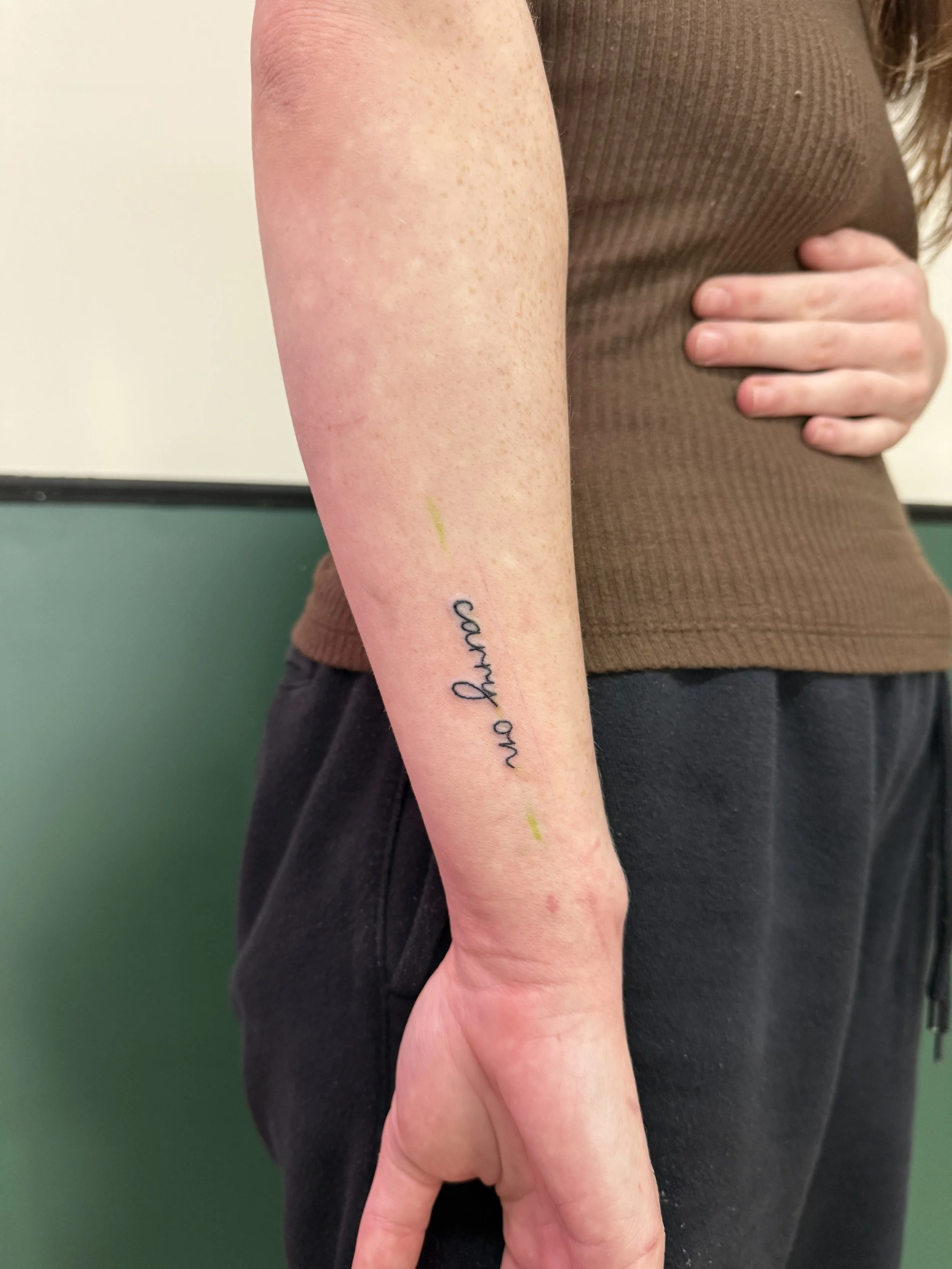 A person's arm with a tattoo that says 'no harm' in black cursive script, resting on their side, with their hand touching their stomach, wearing a brown sleeveless top and black pants, standing against a light background with a green lower section.