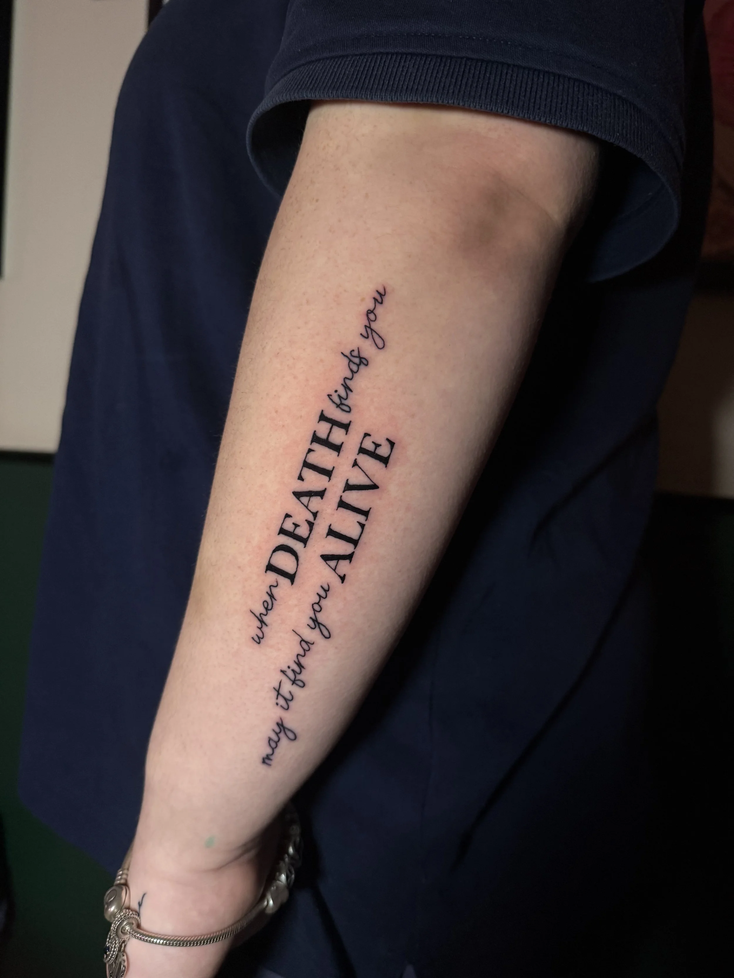 Close-up of a person's forearm with a tattoo that reads, 'I hope when death finds you it finds you alive,' in black ink. The person is wearing a dark-colored short-sleeve shirt, a bracelet, and a ring.