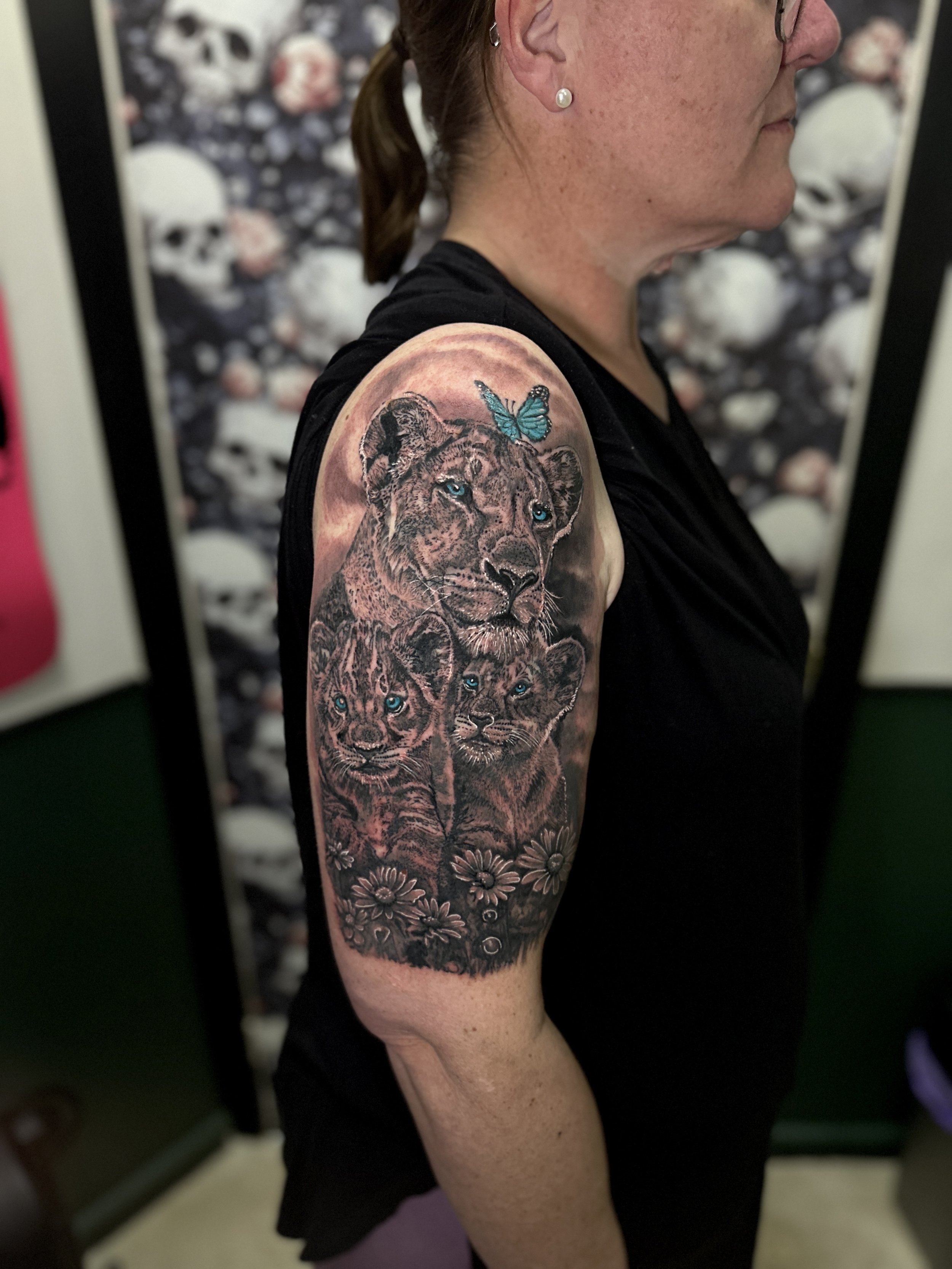 A woman with tan skin and red hair tied back, wearing pearl earrings, displays a detailed tattoo of a lion's face with piercing blue eyes on her shoulder. The tattoo includes a blue butterfly near the lion's ear and surrounded by flowers, with a real