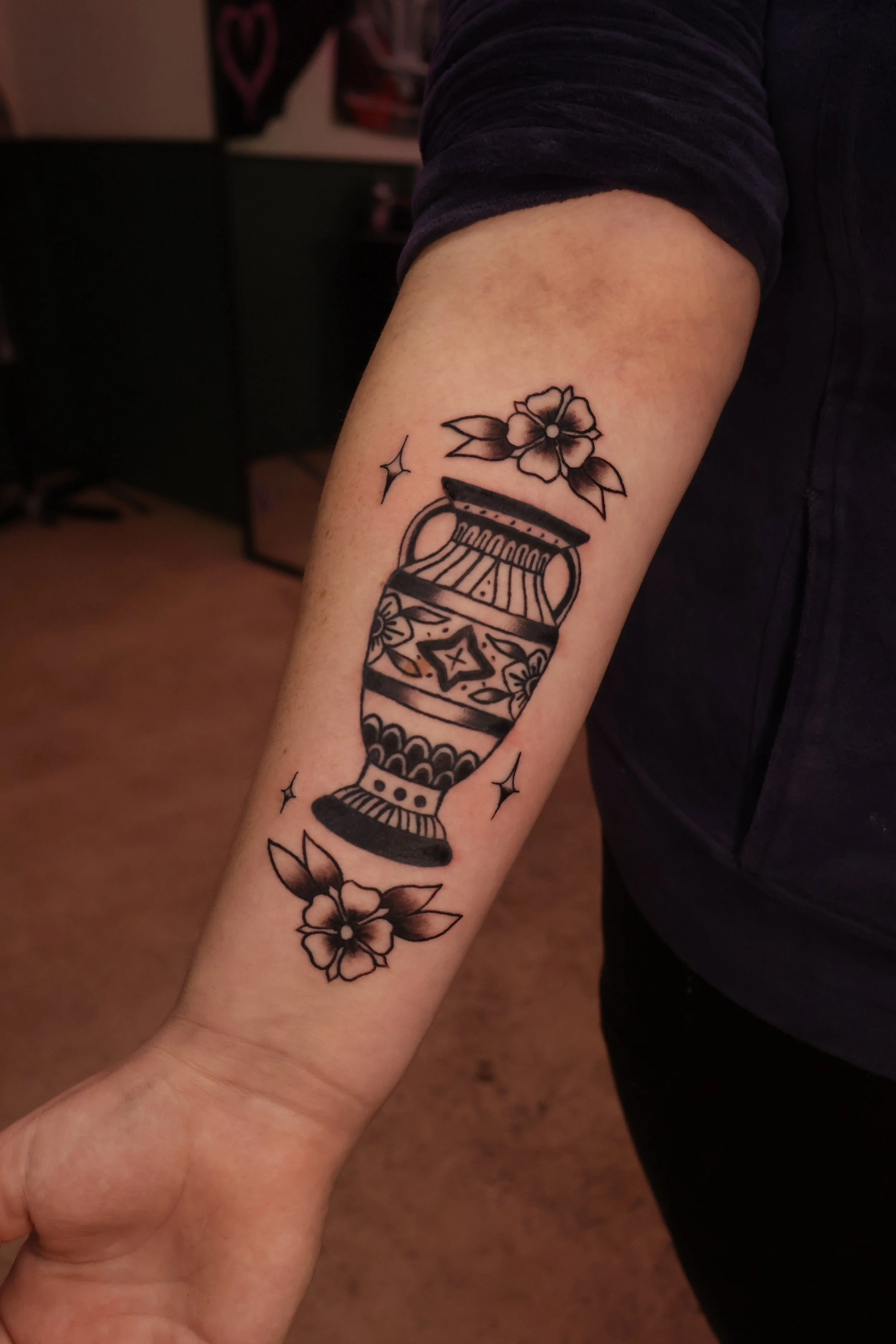 Tattoo of a flower, a star, and a decorative vase on a person's forearm.