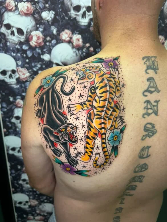 A colorful tattoo of a tiger and a panther surrounded by flowers and stars on a man's shoulder, with a background of skulls and roses.