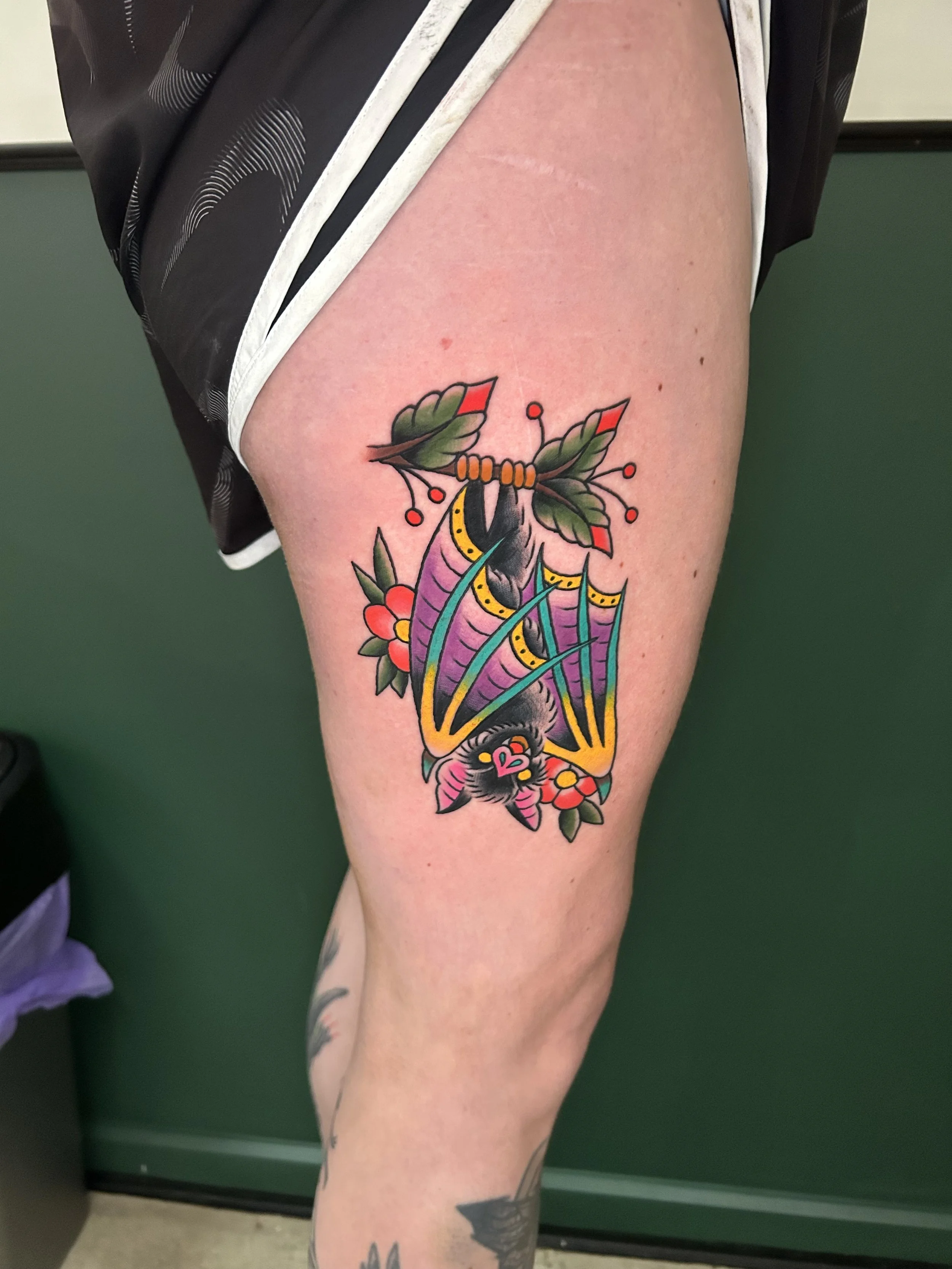 Colorful tattoo of a bat with intricate wing details, surrounded by flowers and leaves on a person's upper thigh.