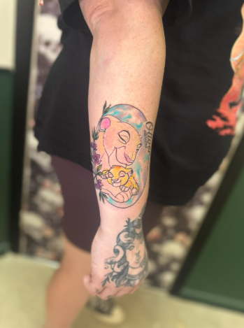 Colorful tattoo of a smiling, moon-faced character with blonde hair and a yellow duck, on a person's arm.