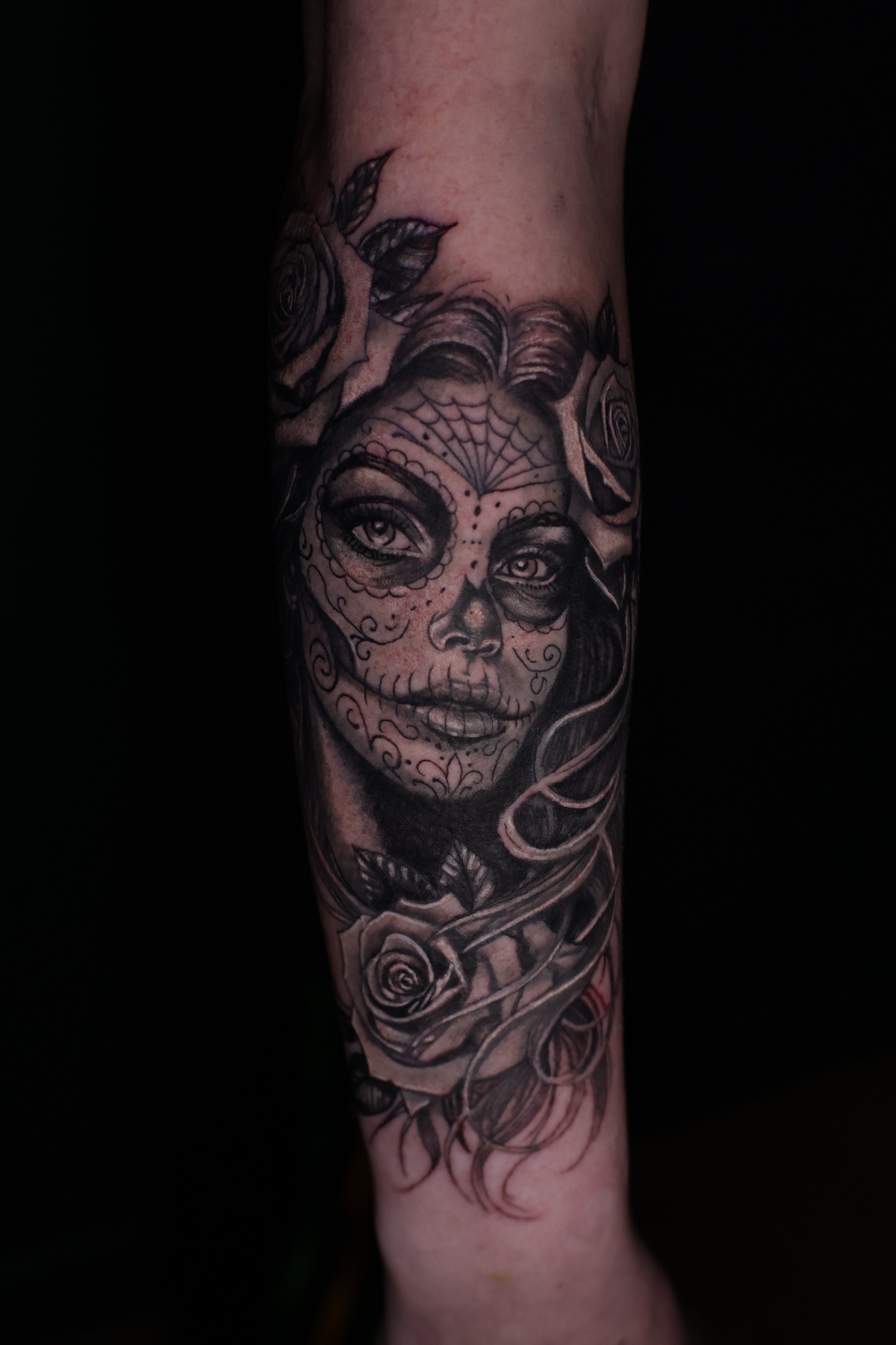 Close-up of a person's arm with a detailed black and grey tattoo featuring a woman's face, roses, and decorative elements.