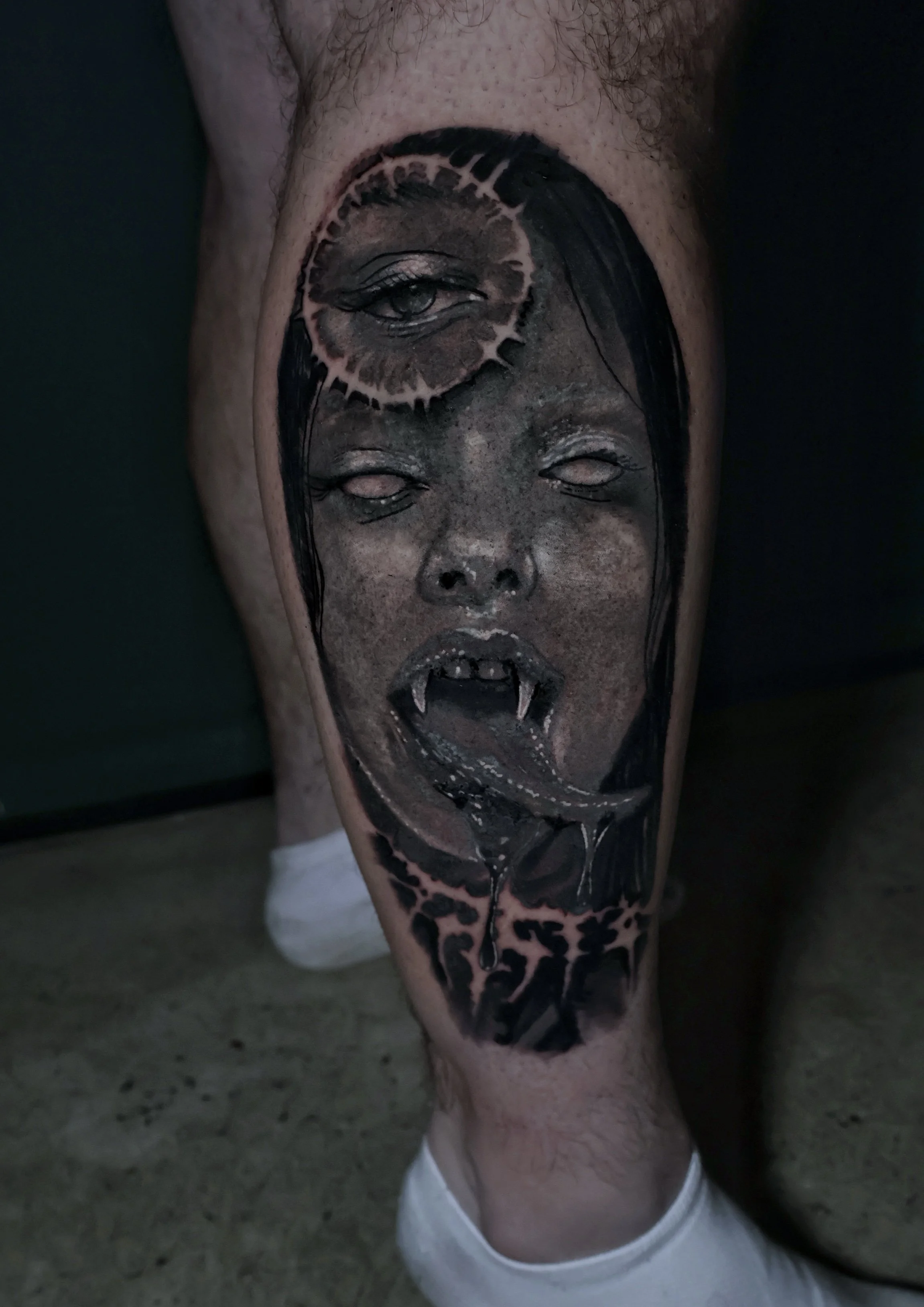 Tattoo on person's leg featuring an eye, a woman's face with open mouth and fangs, with dark coloring and intricate details.