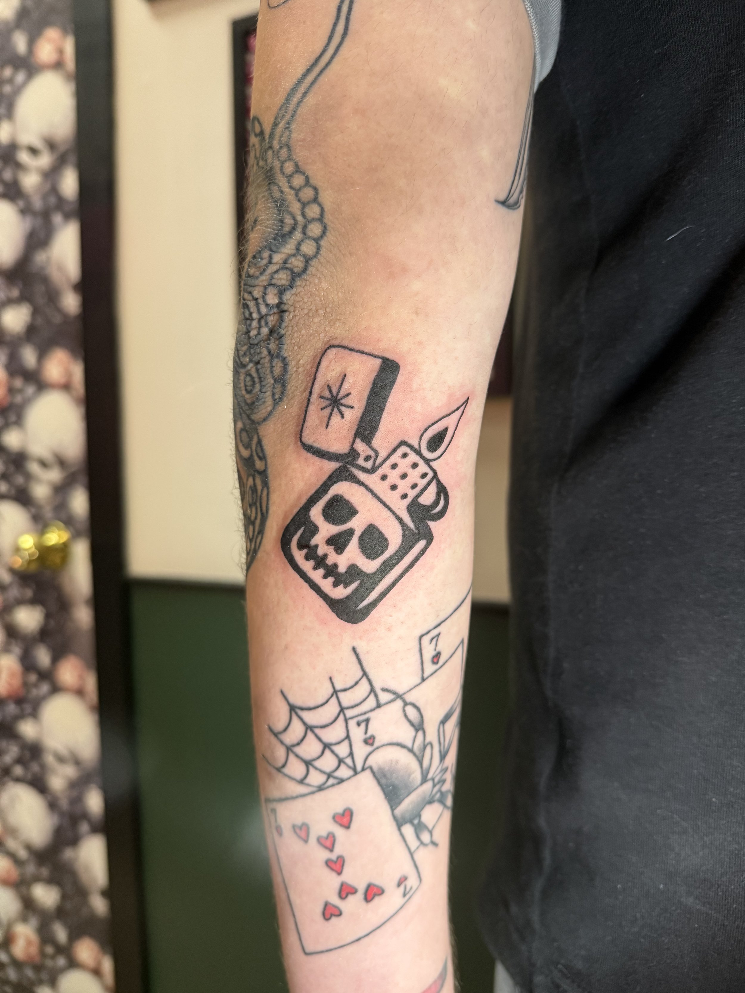 Tattoo of a skull inside a lighter with a spider web below and three red hearts above.