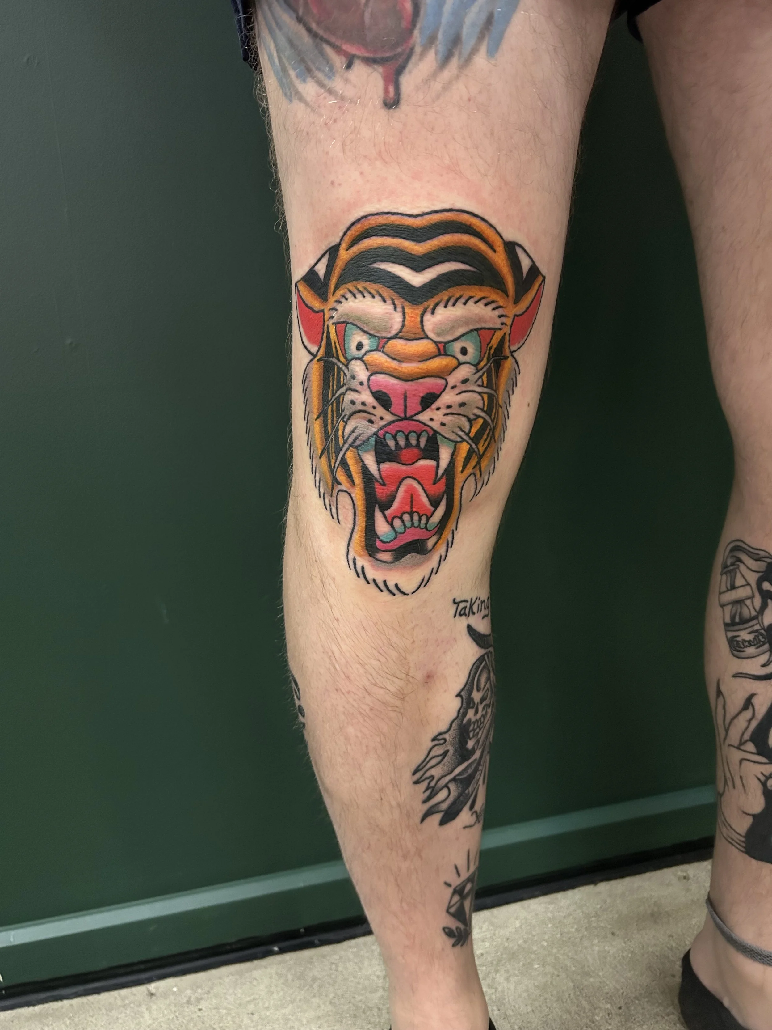 Tattoo of a fierce tiger's face on a person's lower leg, with vibrant colors including orange, white, black, and hints of blue and pink.