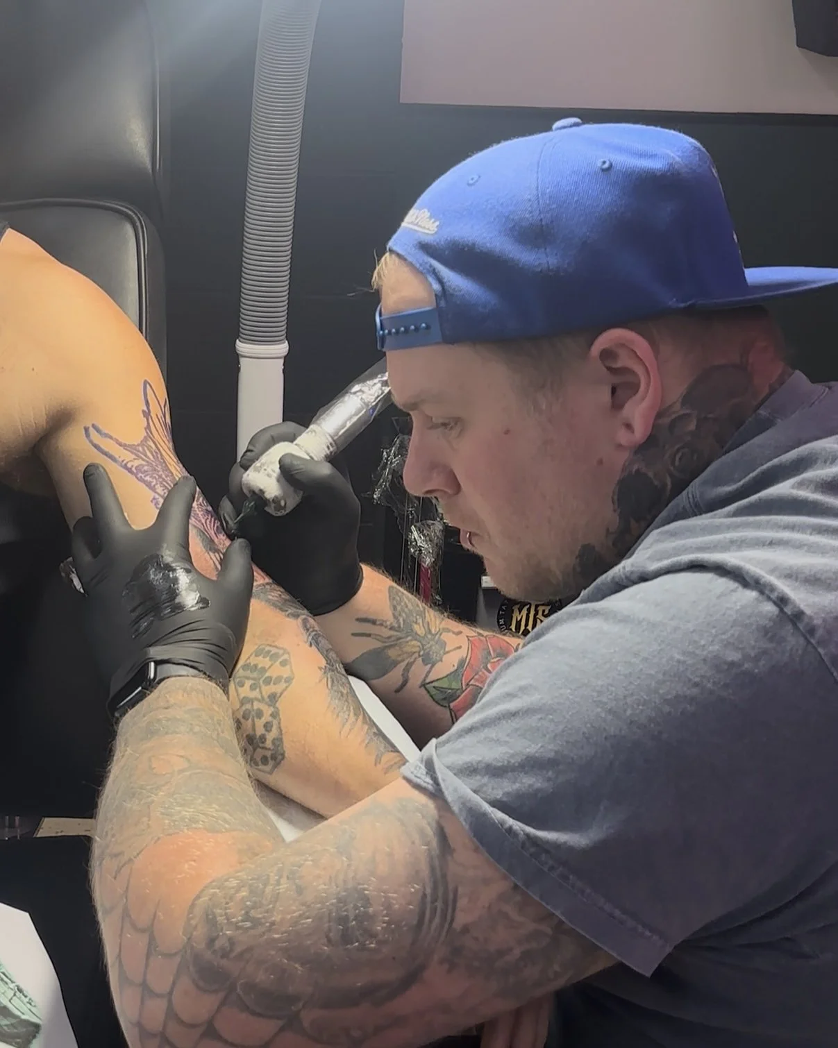 Tattoo artist in a gray t-shirt and blue backward cap tattooing a client's arm, with tattoo equipment.