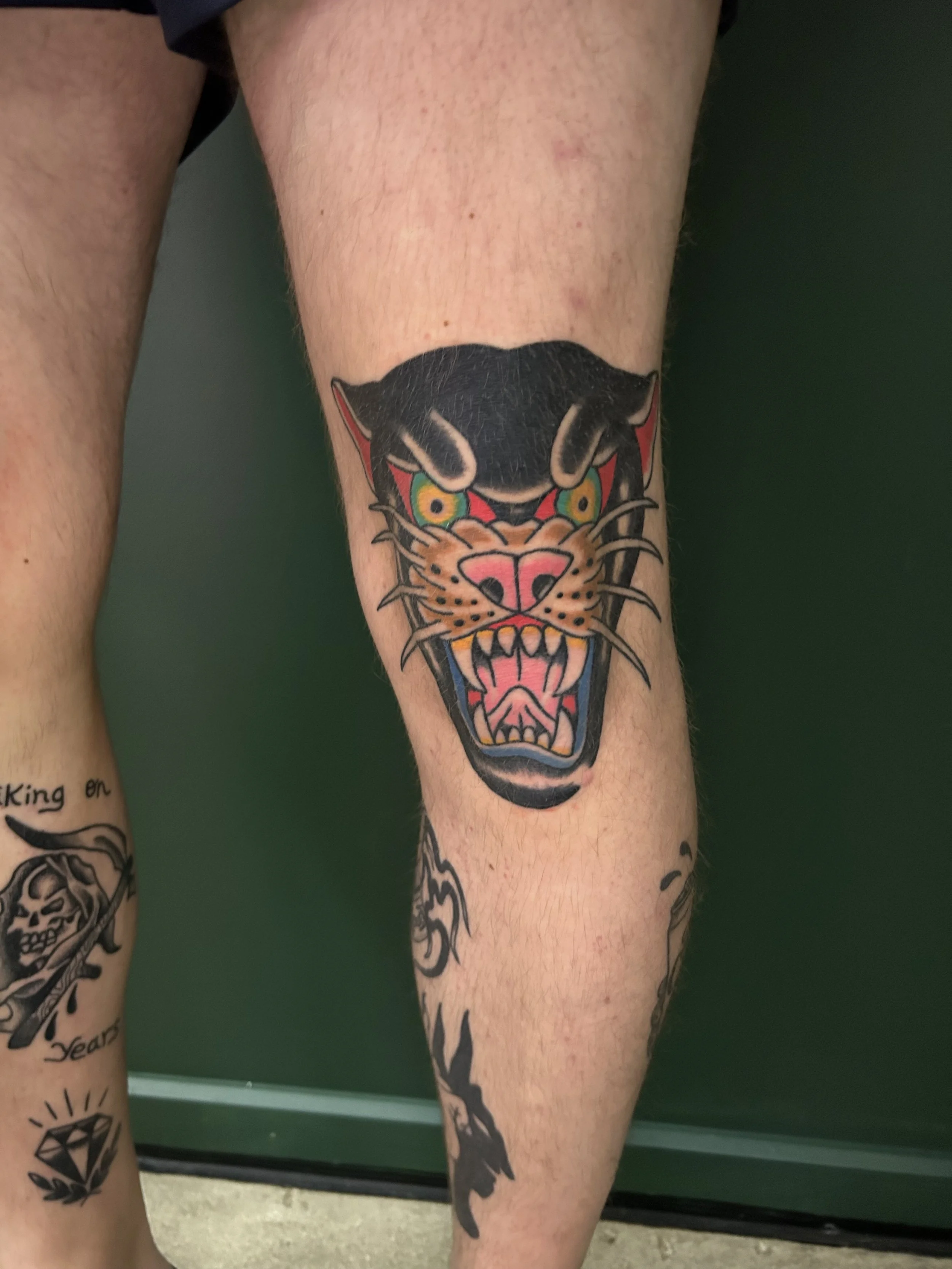 A person's leg with a colorful, traditional-style tattoo of a panther face on the knee. The tattoo features bold black outlines, bright yellow eyes, red accents, and a pink nose.