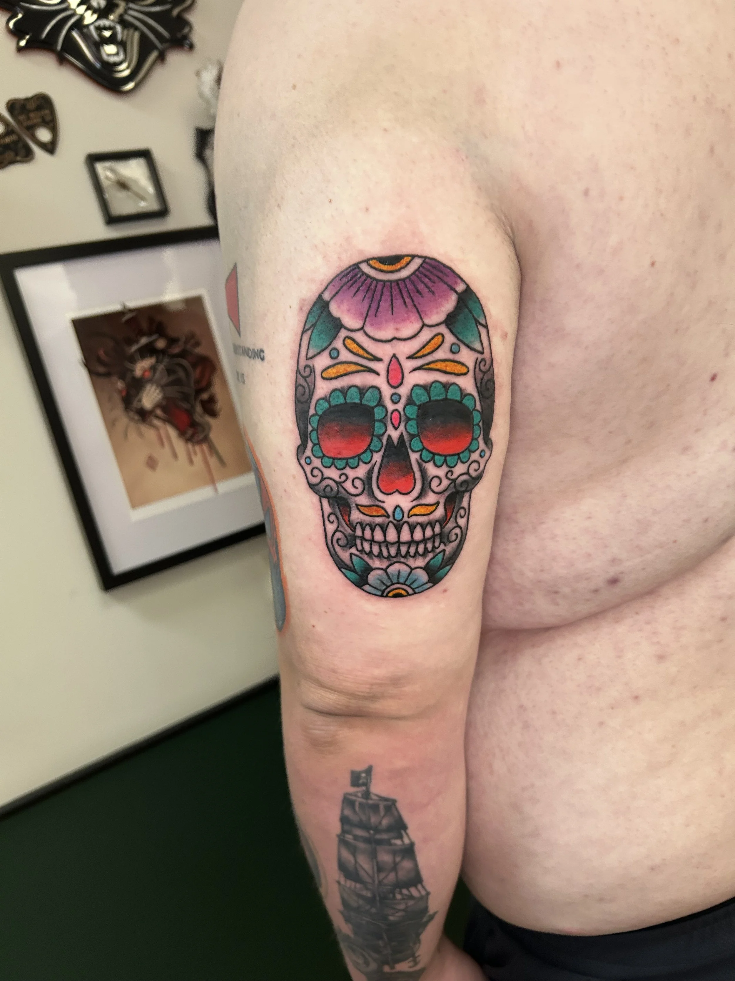 Colorful sugar skull tattoo on person's upper arm, with purple, orange, red, and teal details, and a lighthouse tattoo on lower arm.
