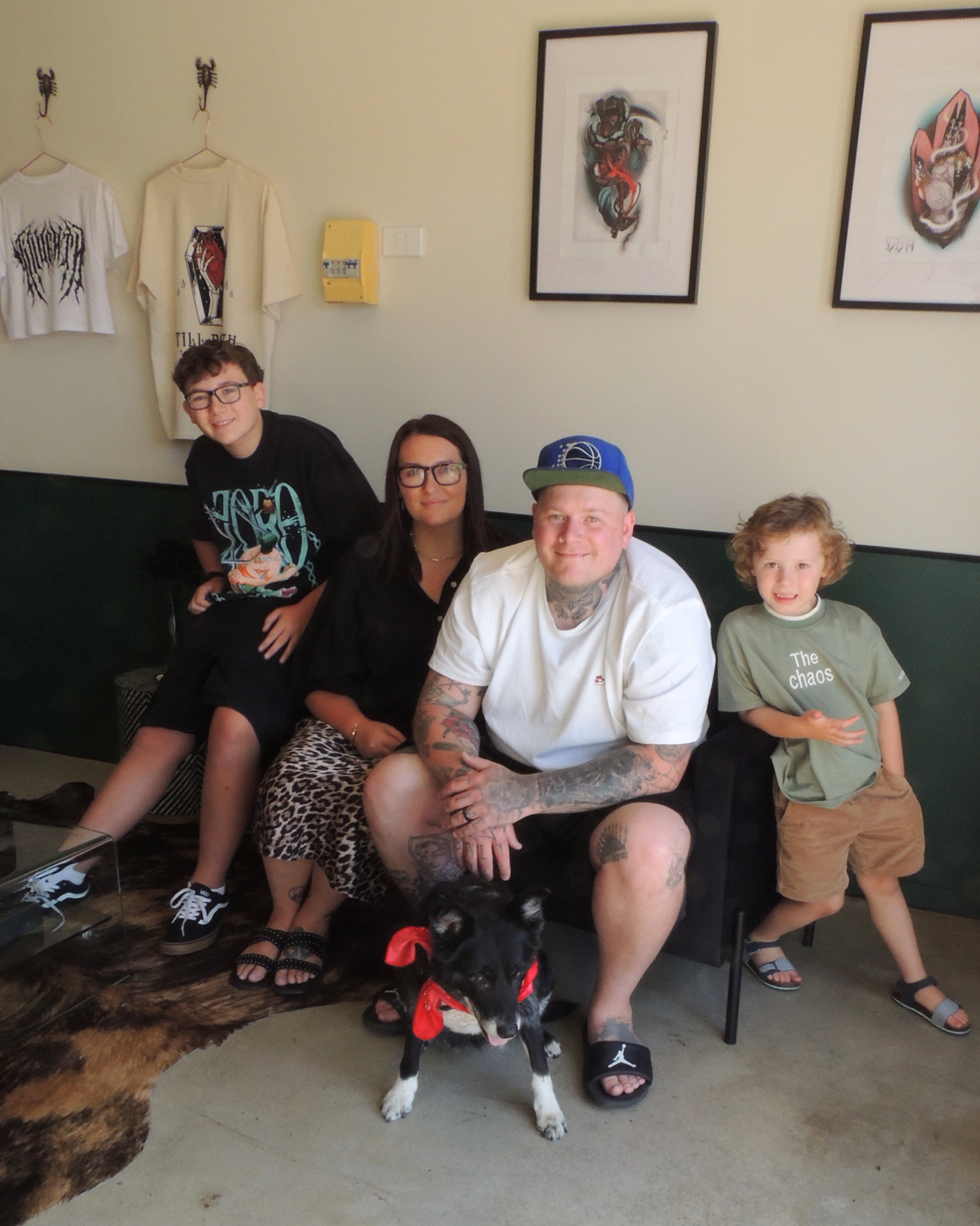 Group of four people and a dog sitting in a room with art on the wall. The dog is a black and white puppy with a red bandana They are smiling and sitting on chairs, with various clothing and accessories visible. The background includes framed artwork and shirts hanging on the wall.