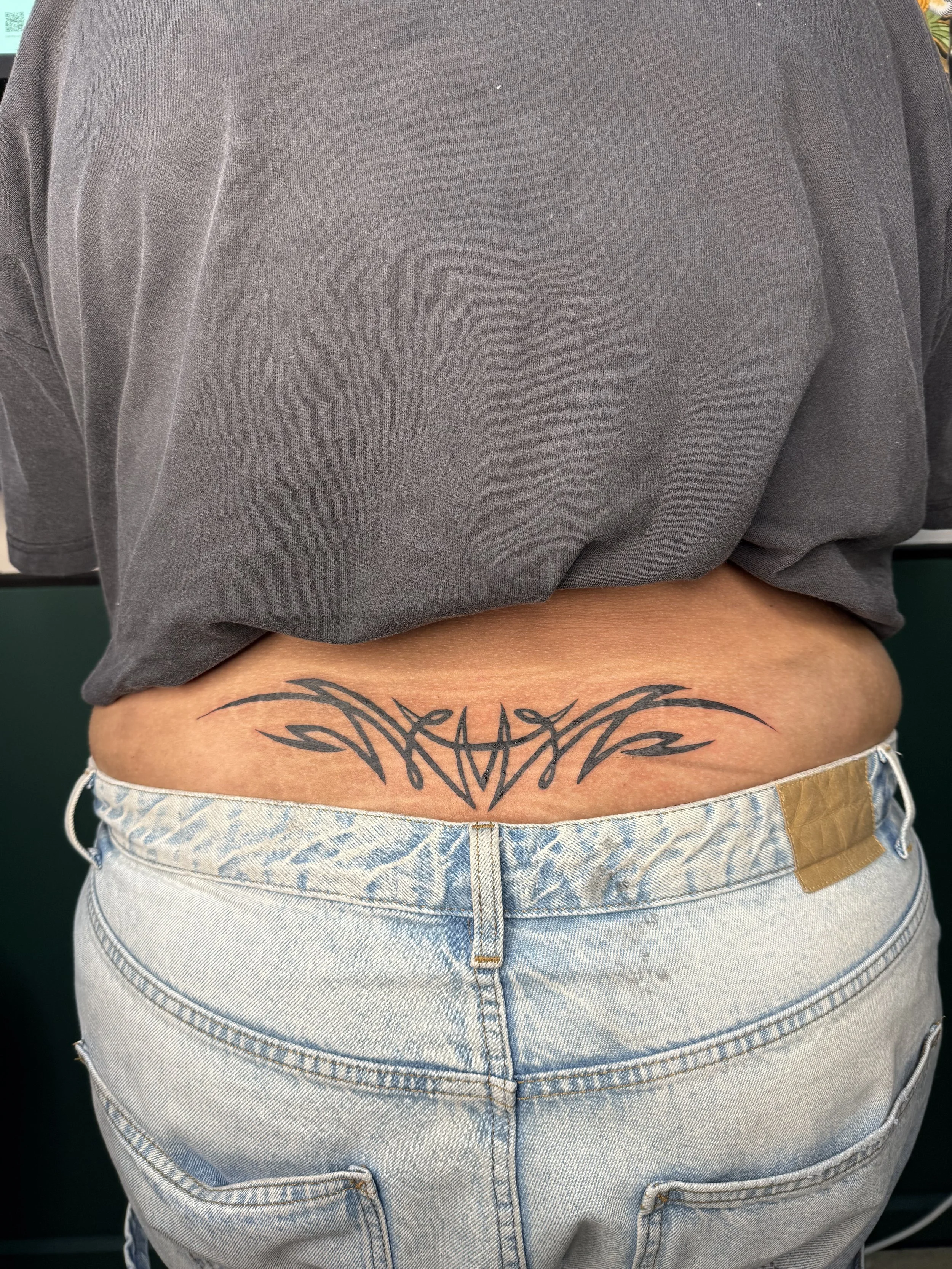 A person's lower back and waist, showing a tattoo on the lower back above the waistband of light denim jeans, with a gray shirt slightly lifted to reveal the tattoo.