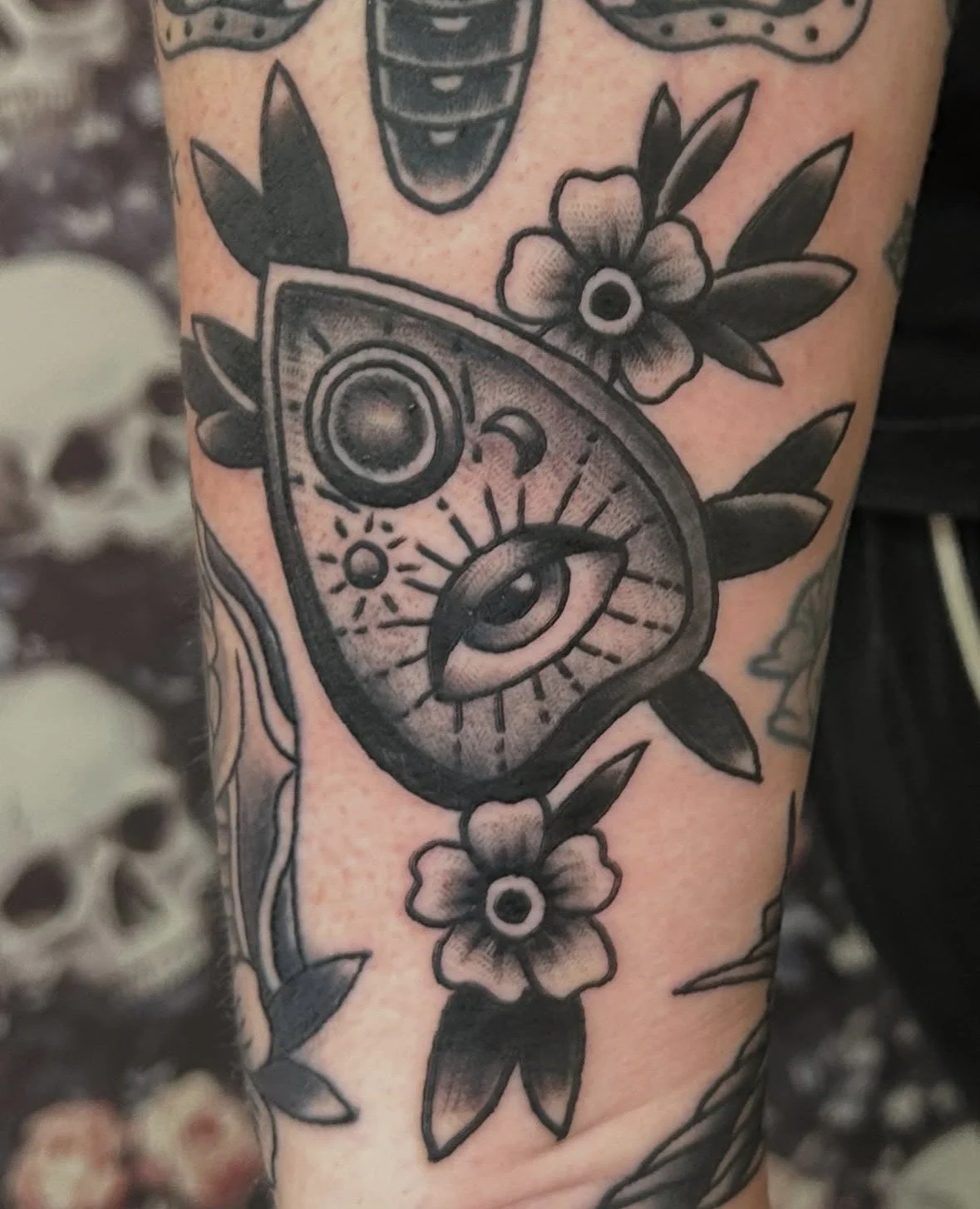 Tattoo of an eye with sun rays, moon, and geometric designs on skin, adorned with flowers and leaves.