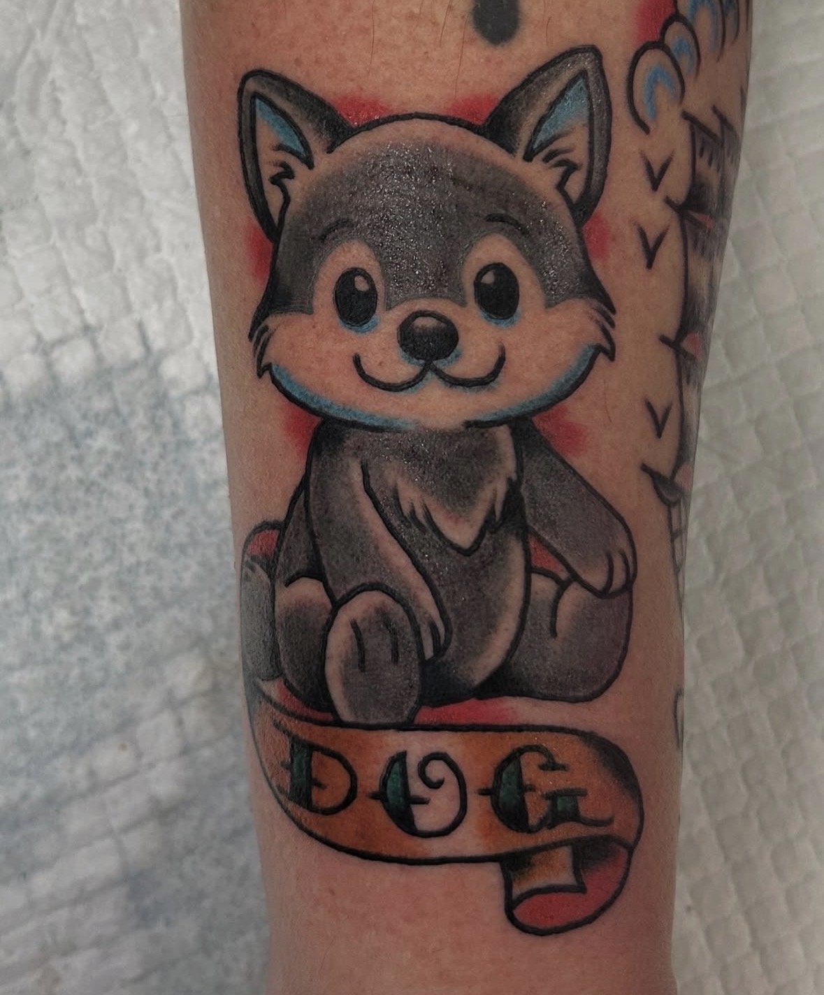 A colorful tattoo of a cute, cartoon-style dog with a gray and black fur pattern, large shiny eyes, a smiling face, and a winking eye. The dog has a blue outline and shading, and is sitting with a playful expression. The tattoo includes a banner unde