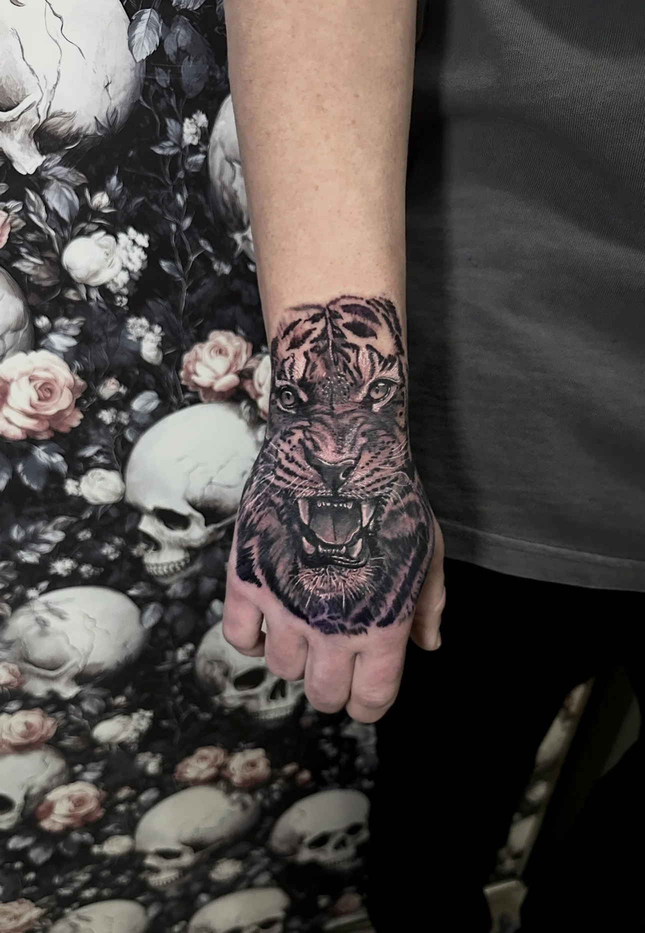 Tattoo of a fierce lion's face on a person's forearm with dark and pinkish colors, background filled with skulls and roses.