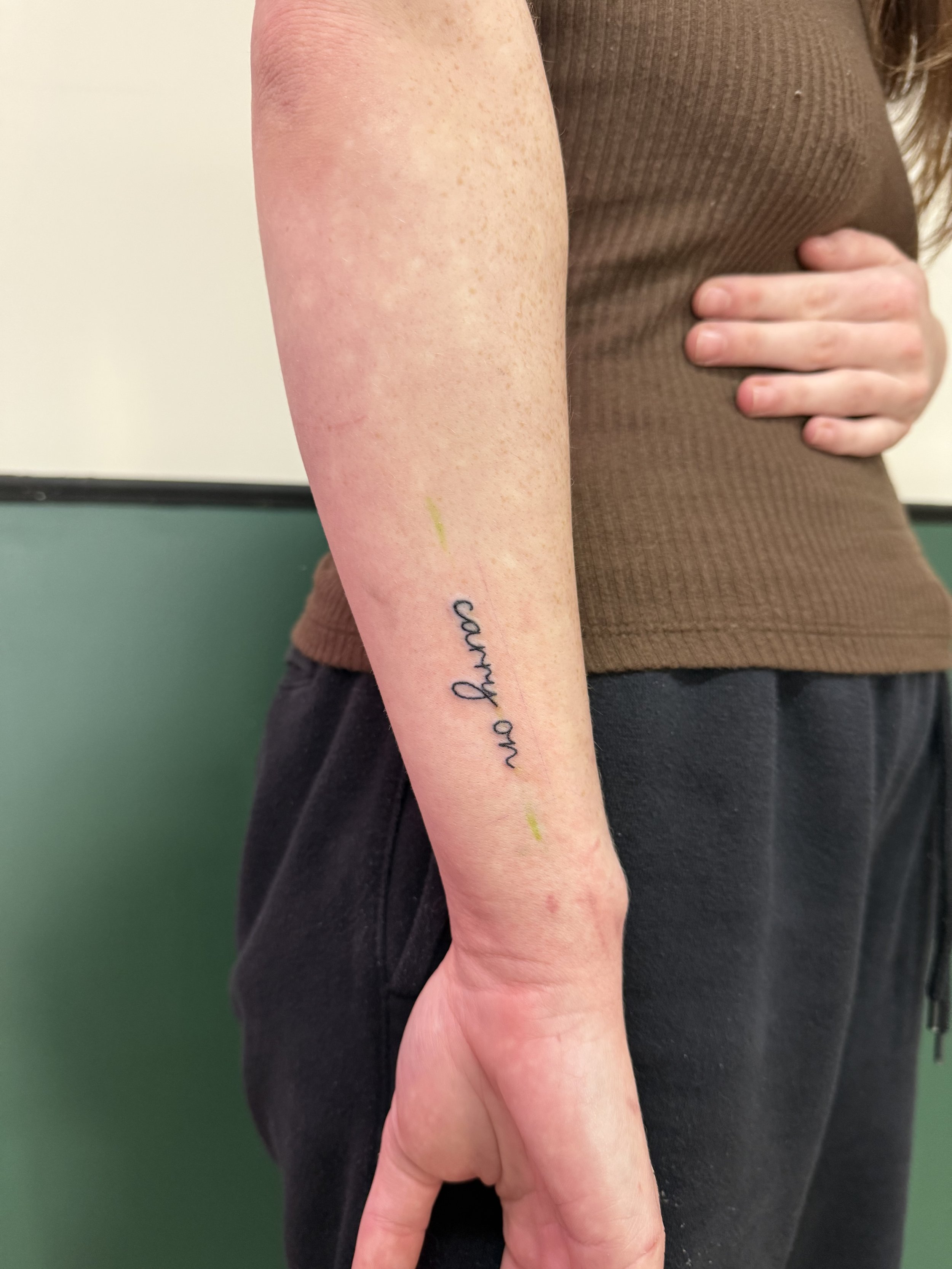 A close-up of a person's left forearm, showing some freckles, a tattoo of black cursive writing, and a yellow mark near the tattoo, with the person's hand resting on their stomach in front of a brown shirt and black pants.