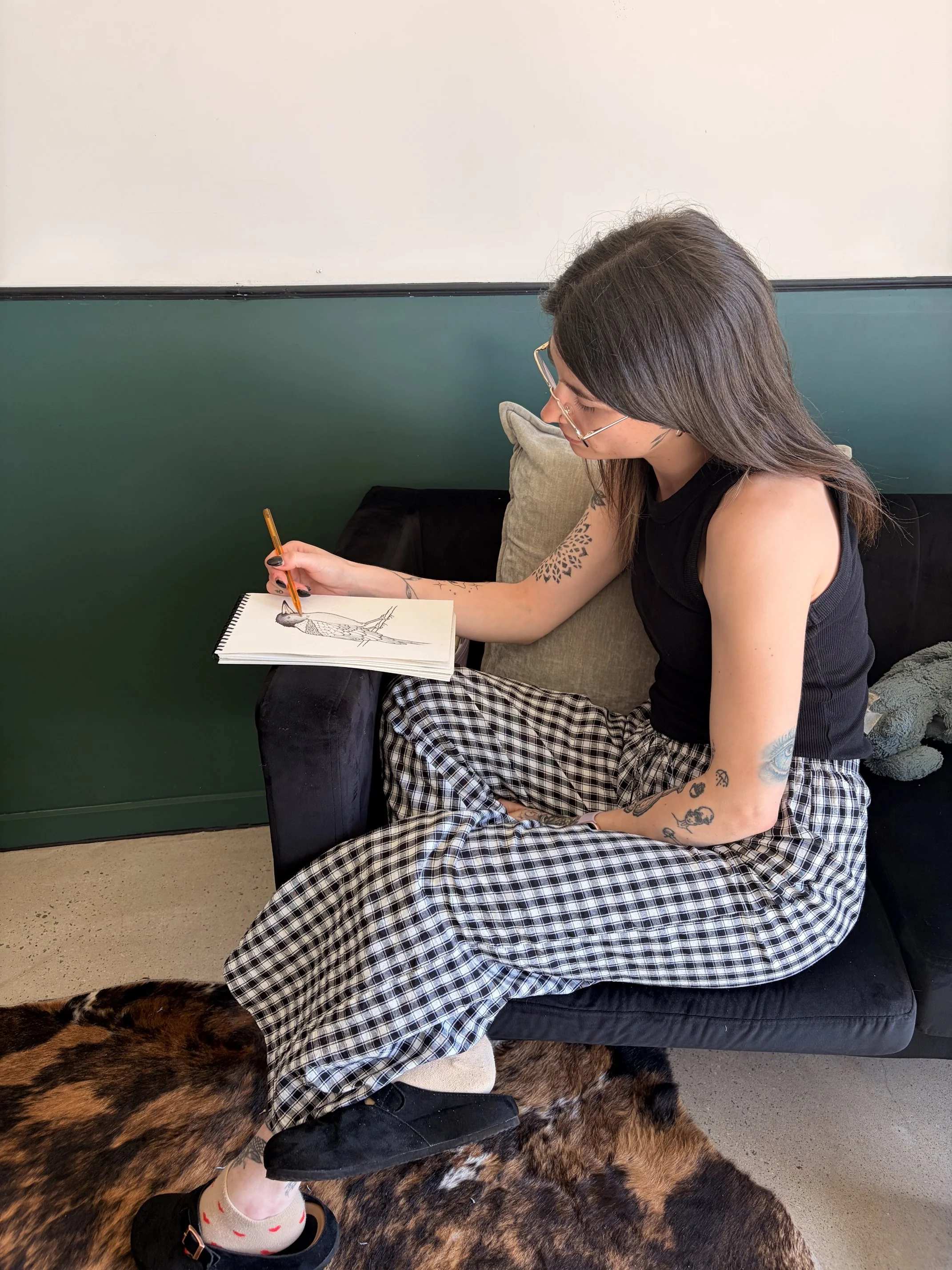 A woman with tattoos on her arms sitting on a black bench, drawing in a sketchbook. She is wearing a black tank top, plaid pants, and black sandals with heart-patterned socks.  The room has a green and white wall with a black trim.