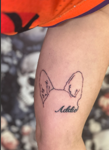 Tattoo of a dogs ear outline with the name 'Addie' written below on a person's thigh.