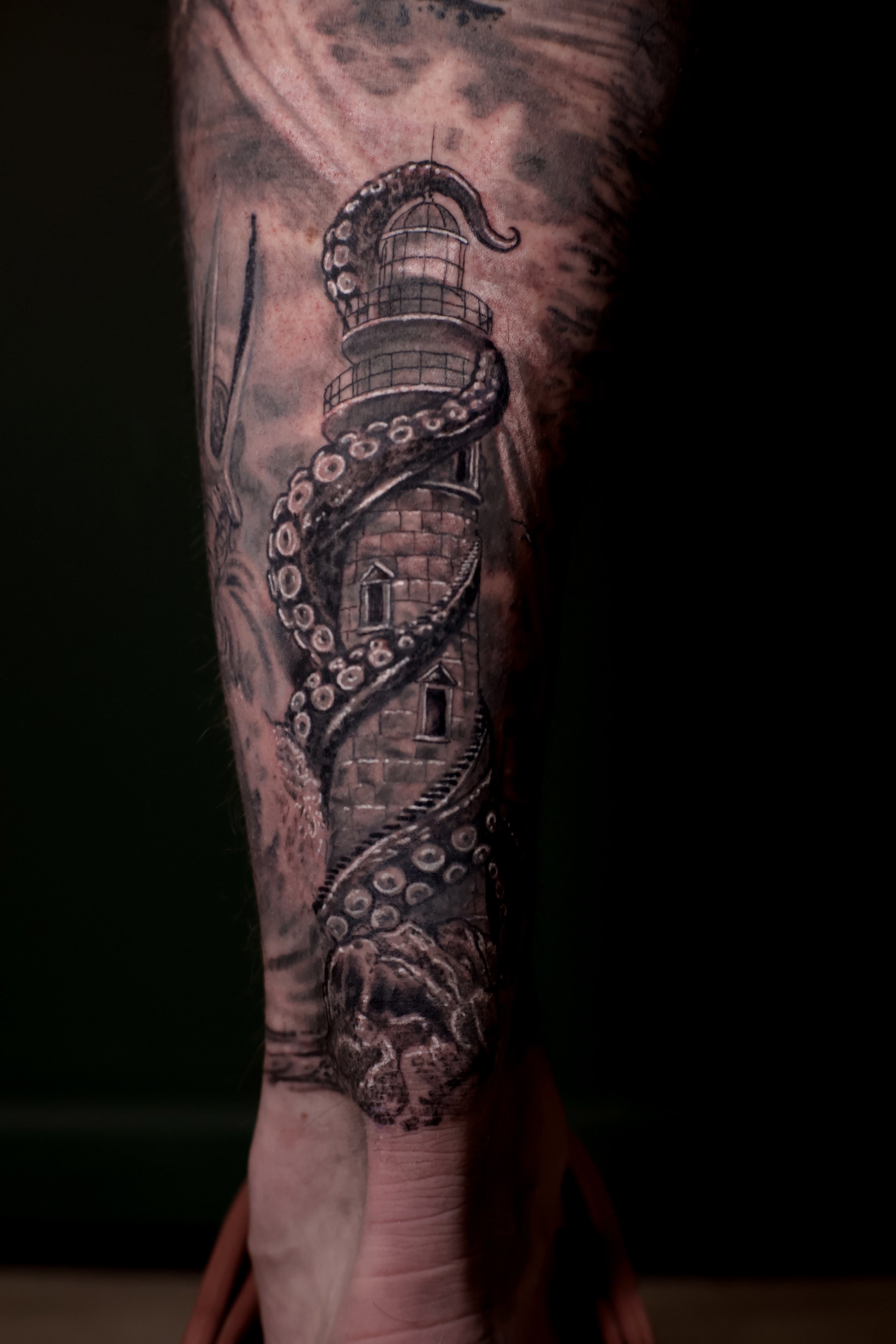 Tattoo of a lighthouse with an octopus's tentacle wrapped around it on a person's leg.