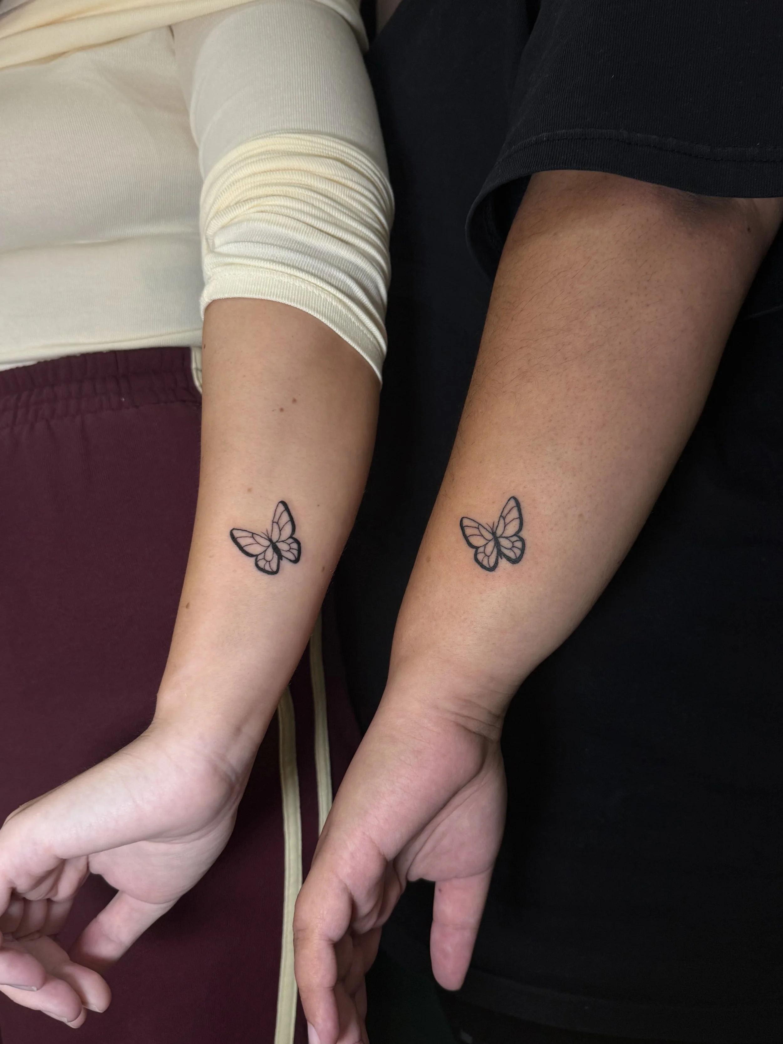Close-up of two people sitting side by side, showing their intertwined hands and forearms, each with a butterfly tattoo on the inner wrist.