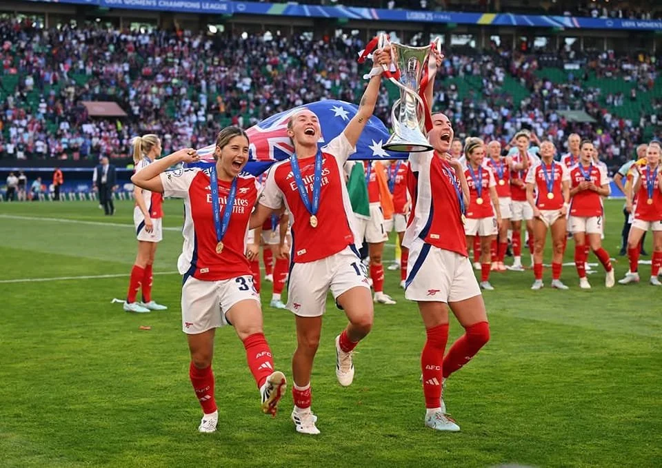 Women soccer players celebrating with a trophy on a stadium field, some wearing medals and cheering.