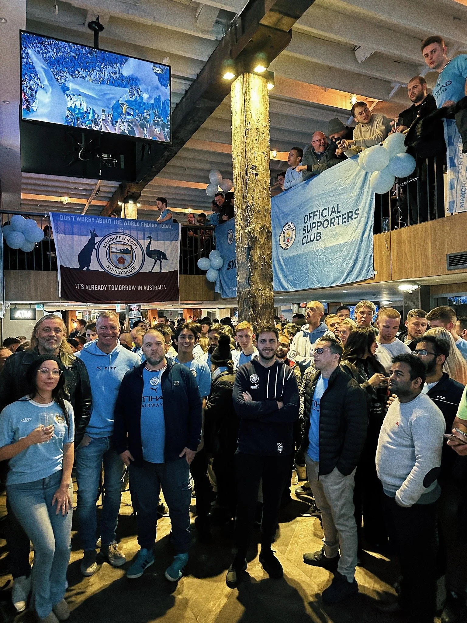 A large group of Manchester City soccer fans gathering in a crowded indoor venue, some wearing team jerseys, with banners and balloons supporting the team. There are people watching a game or event on a screen overhead.