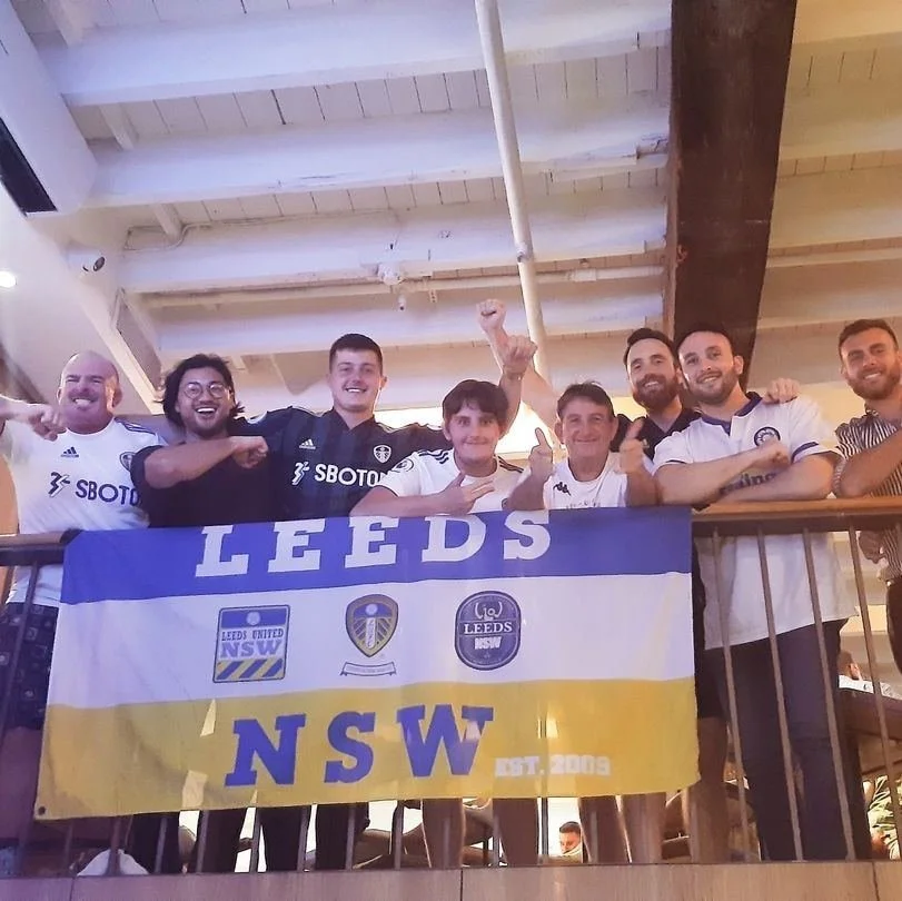 Group of men celebrating behind a Leeds United flag, wearing team jerseys, inside a building with a wooden ceiling.