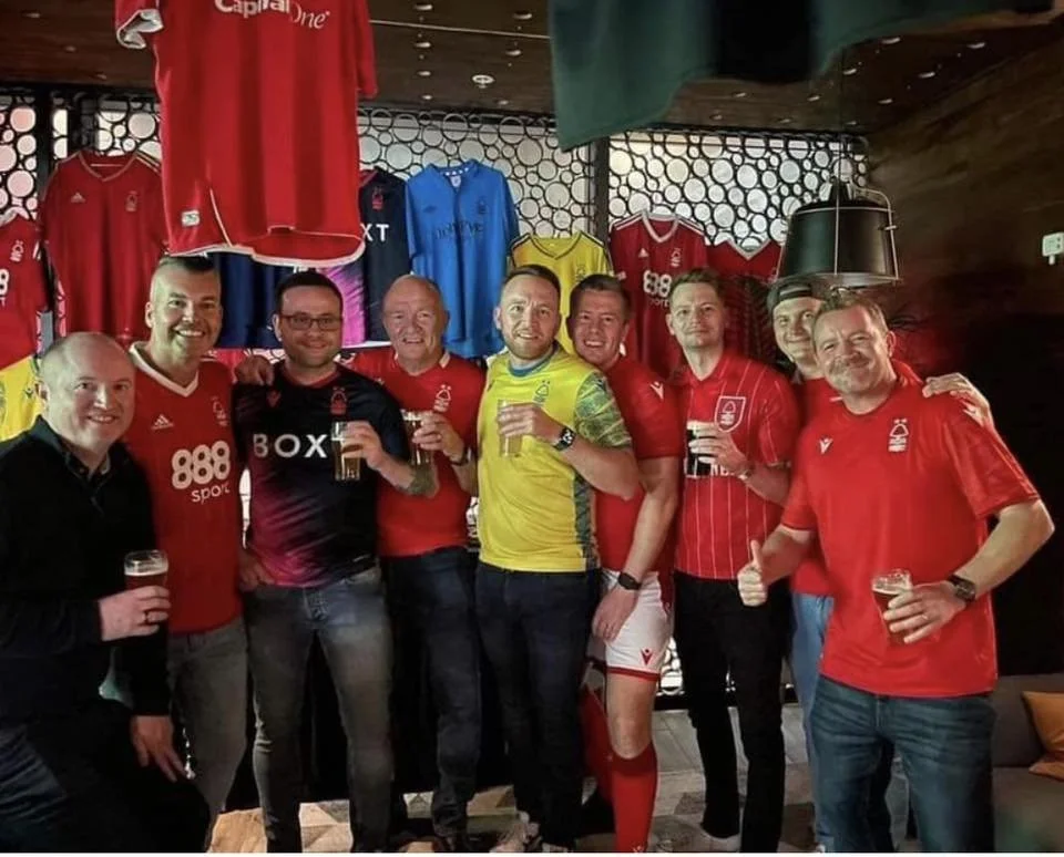 Group of men celebrating with drinks in a sports-themed pub, wearing football jerseys, in front of a wall decorated with various soccer jerseys.
