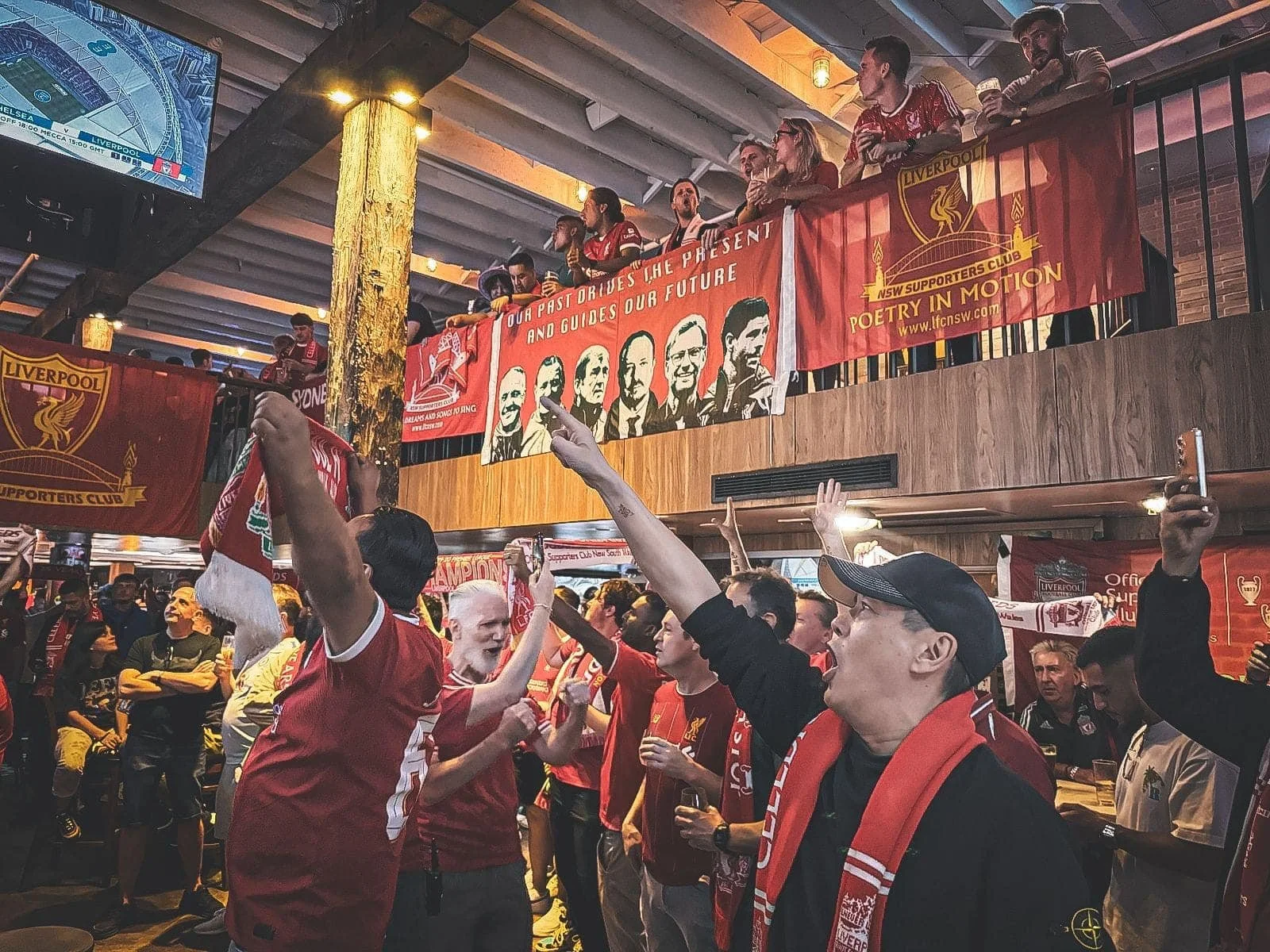 Crowd of Liverpool Football Club fans at Anfield stadium waving scarves and holding banners in red and white, with one prominent banner reading 'Anfield Down Under' and the club's emblem.