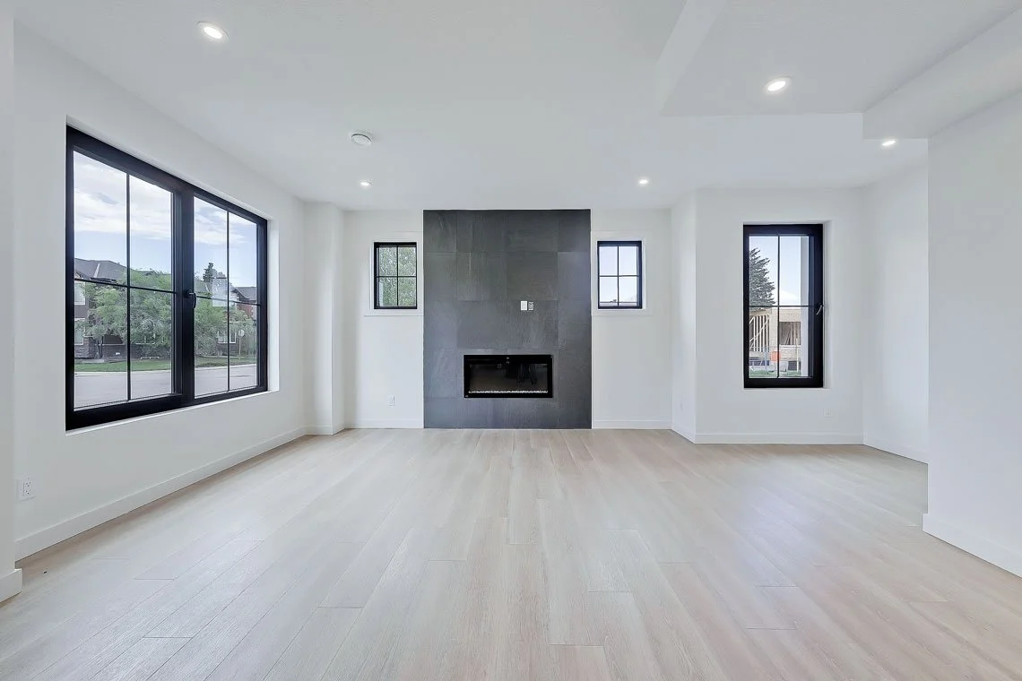 Empty living room with large windows, a black fireplace, and light wood flooring.
