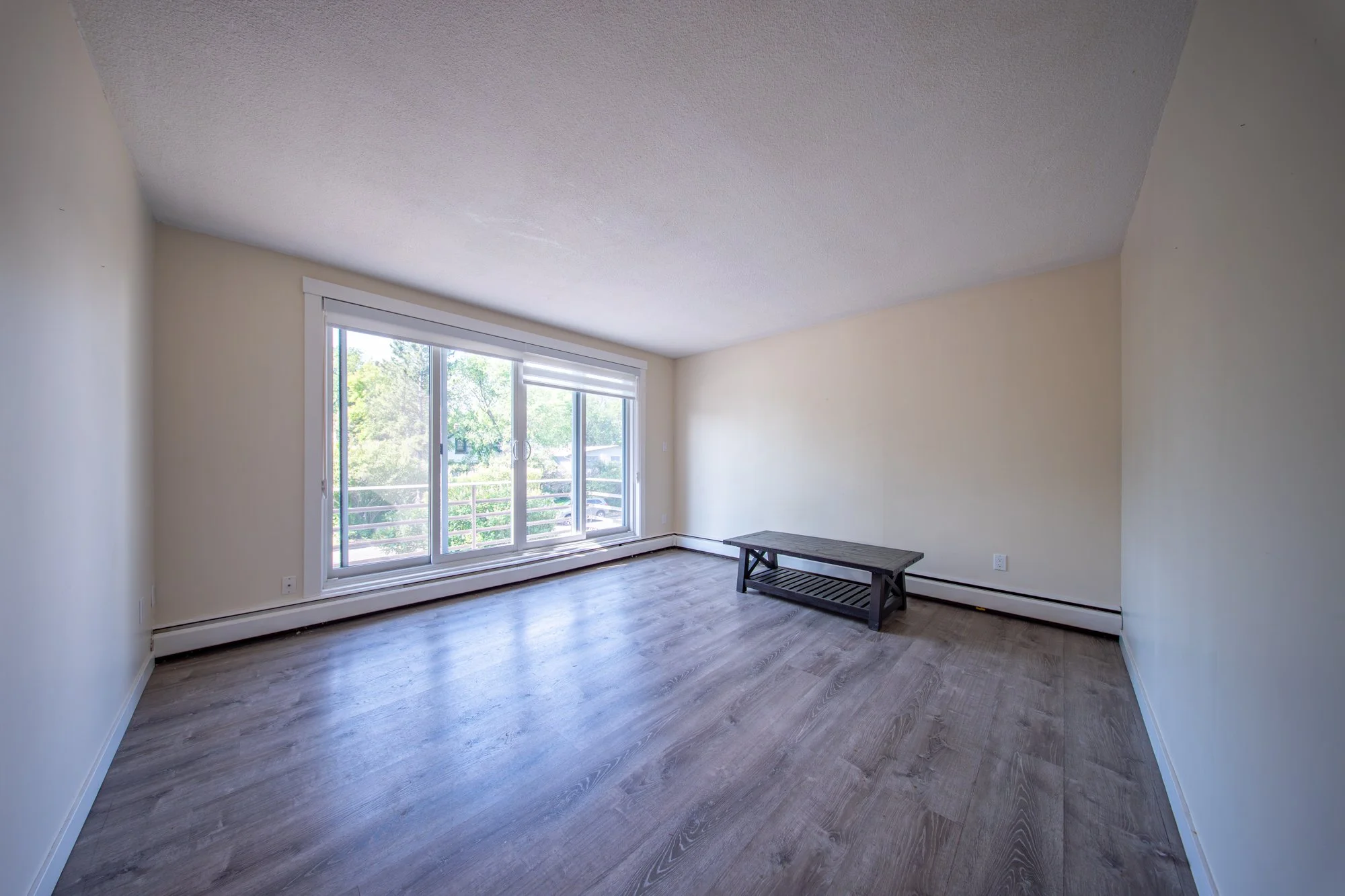 Empty living room with large sliding glass door, light-colored walls, wood-like flooring, and a small wooden bench.