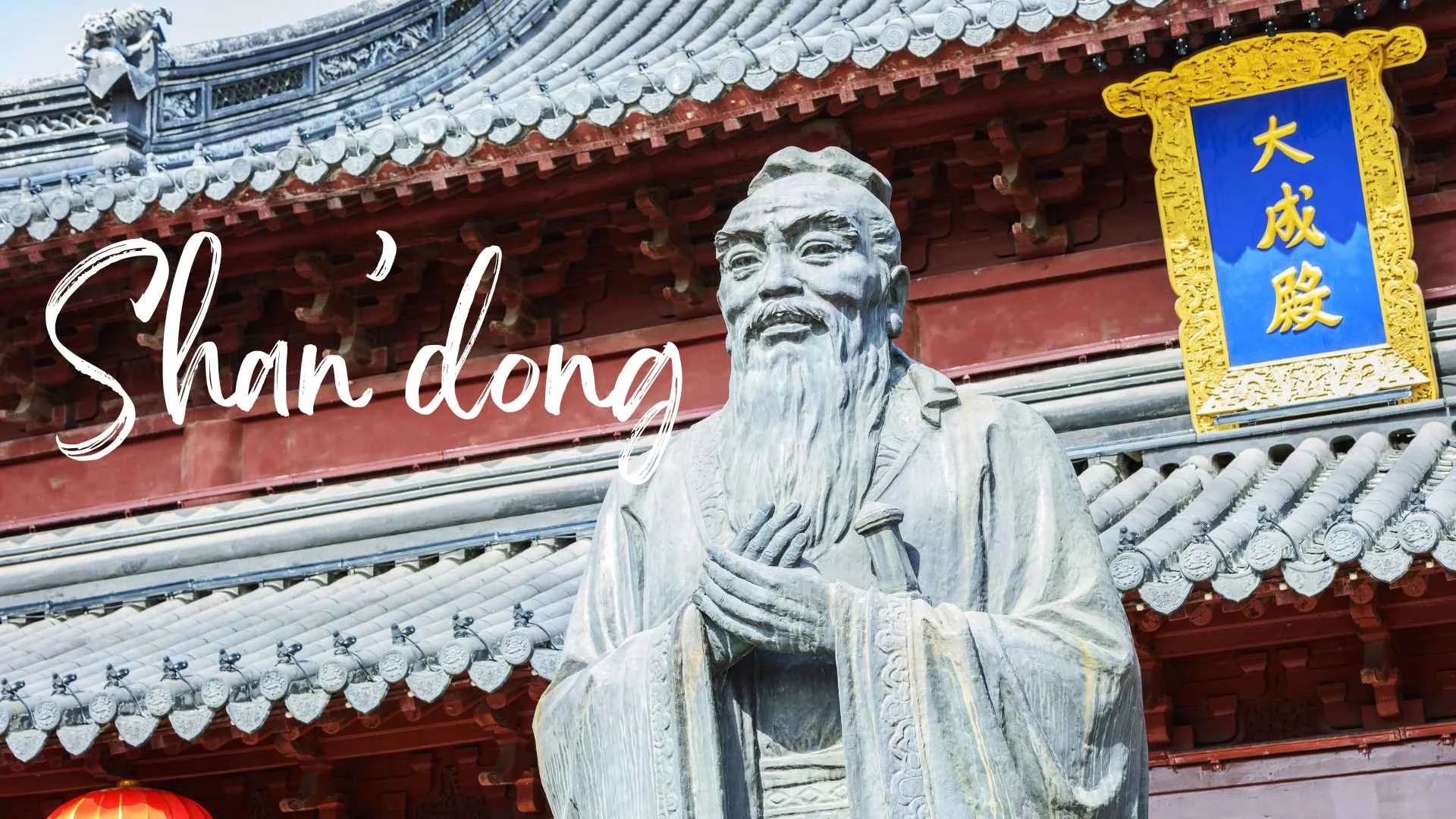 Statue of a bearded Asian man holding a scroll or document in front of a traditional Chinese building with gray tiled roof, red walls, blue and yellow sign with Chinese characters, and paper lanterns.