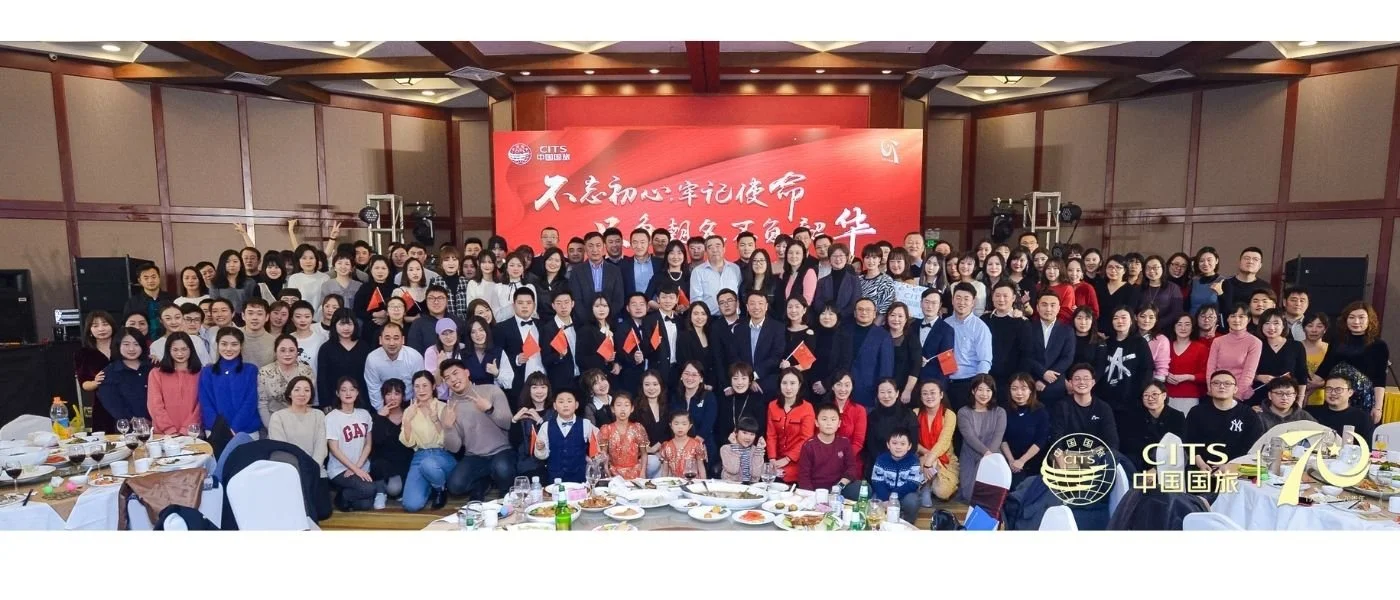 A large group of people gathered in a banquet hall, posing for a group photo with a red banner in the background. The table in front has dishes and drinks, indicating a celebration or event.