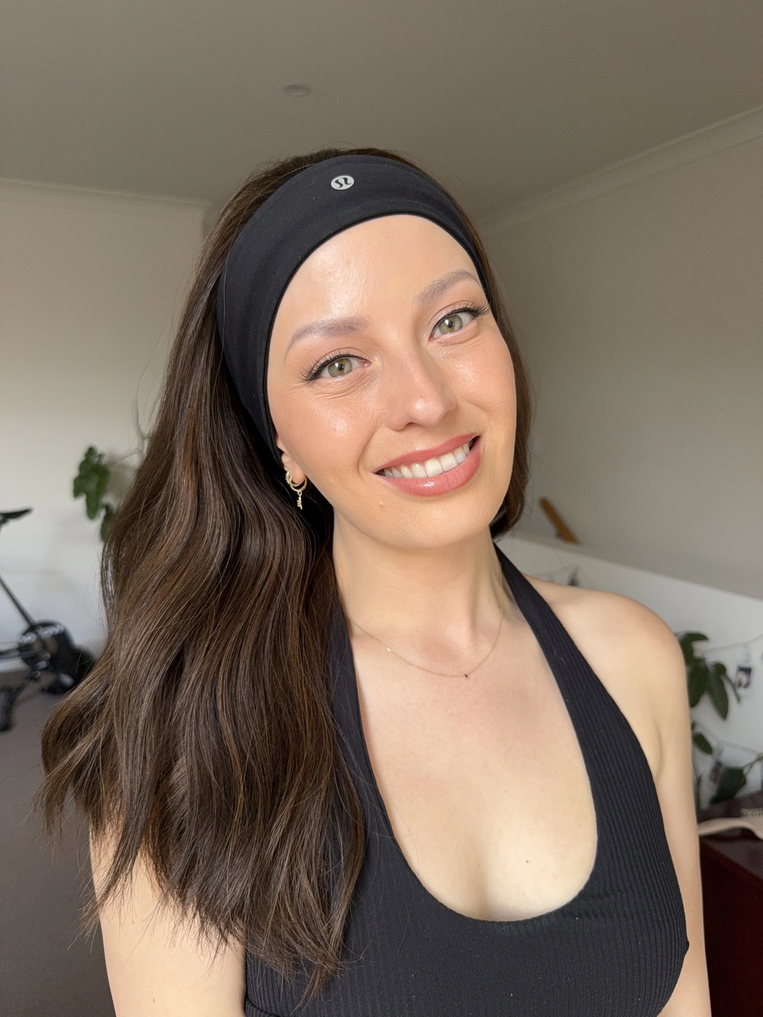 A woman with long brown hair, wearing a black headband and black tank top, smiling indoors.