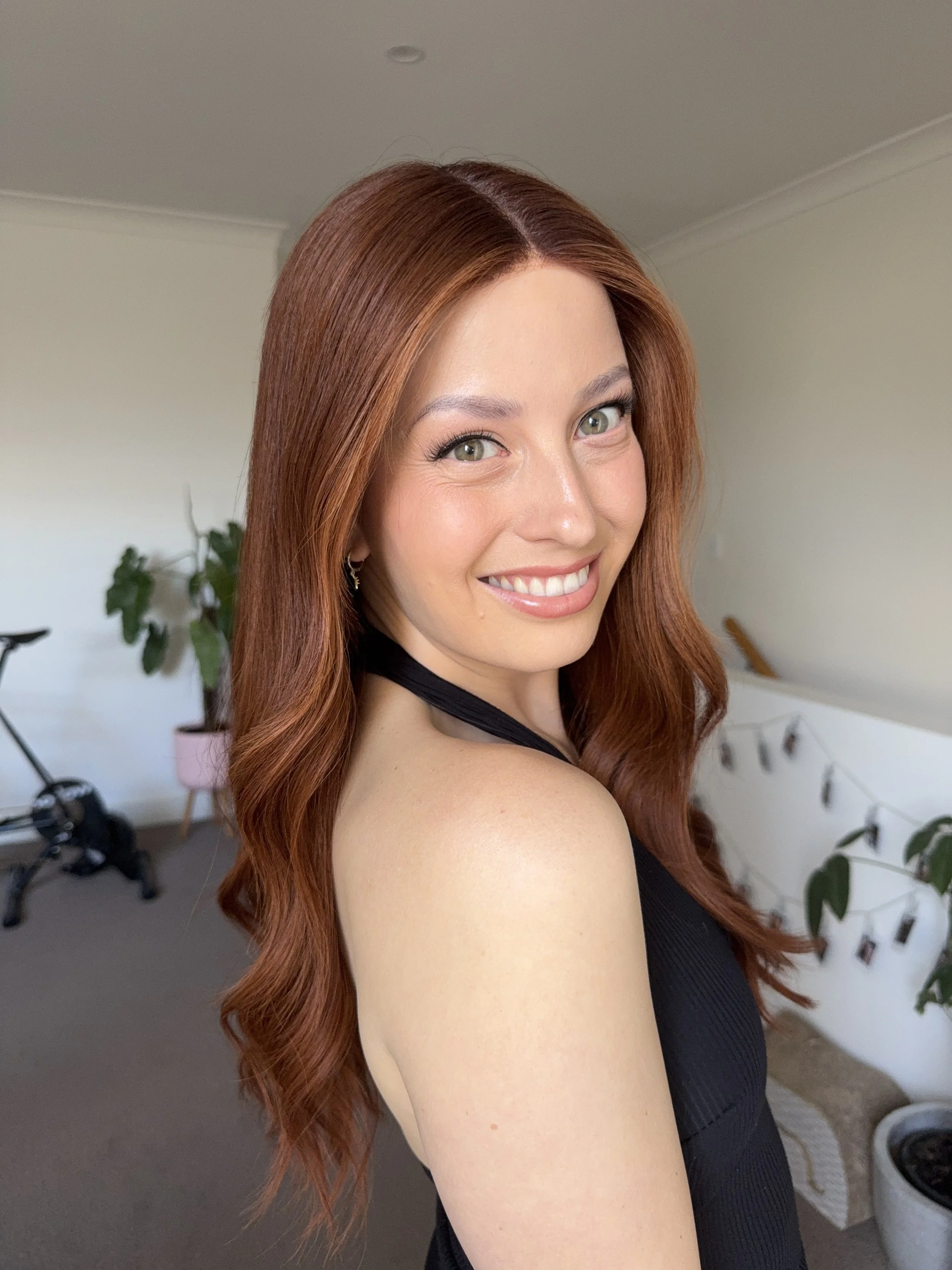 A woman with long, wavy auburn hair smiling at the camera, wearing a black sleeveless top, inside a room with plants and a string of Polaroid photos hanging on the wall.