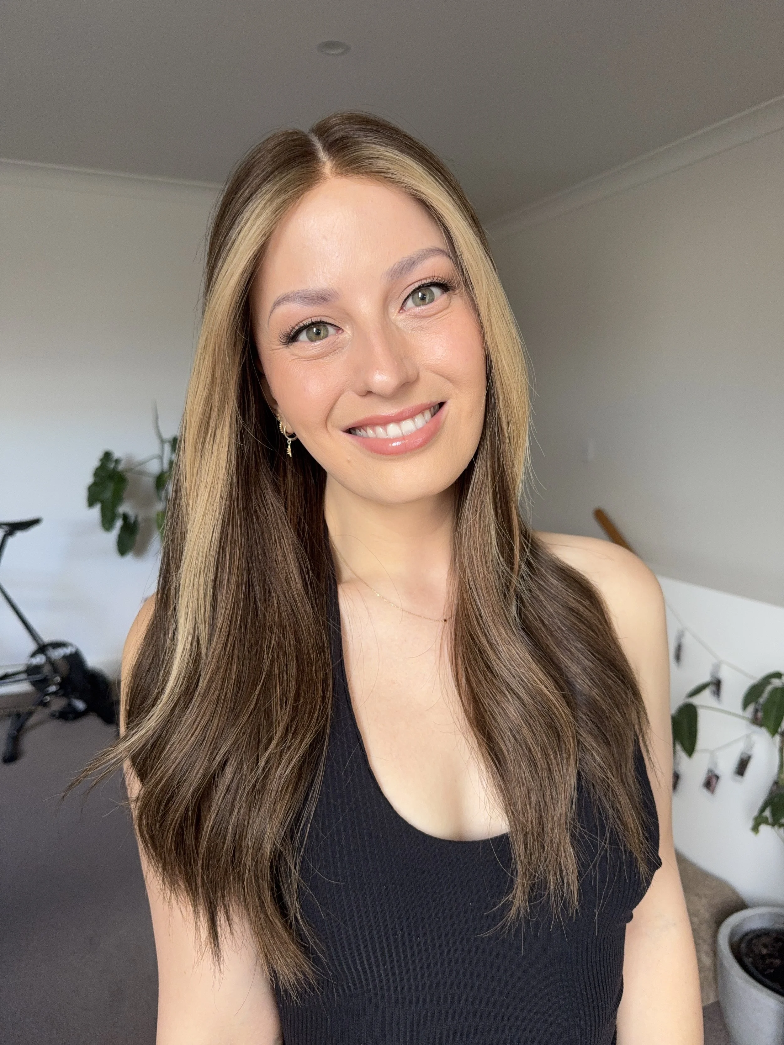 A woman with long brown hair and light makeup, smiling at the camera, wearing a black sleeveless top, with a blurred background including a bike, plants, and a wall with photo clips.