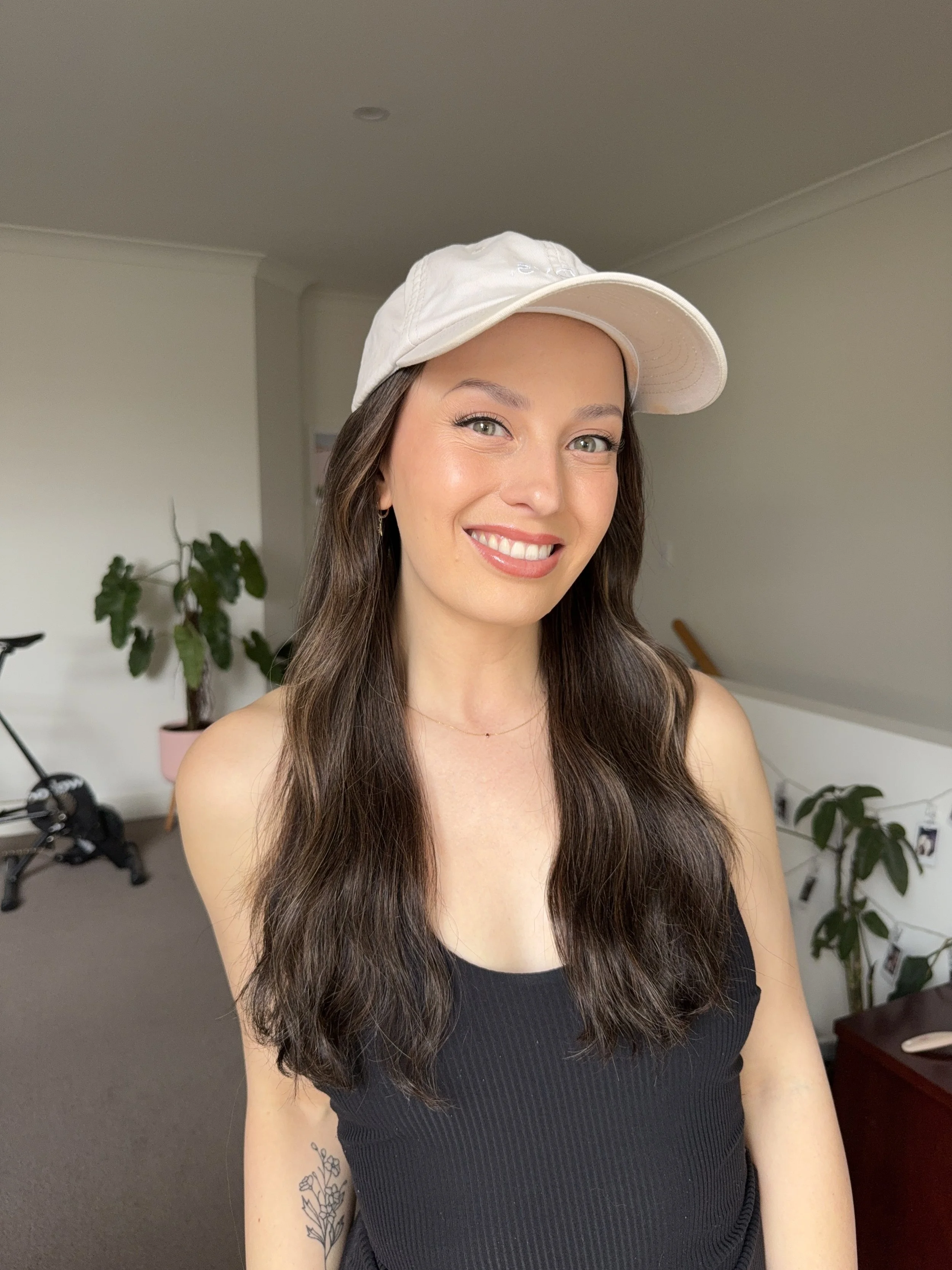 Young woman with long brown hair, wearing a white cap and black tank top, smiling in a room with potted plants and a photo wall.