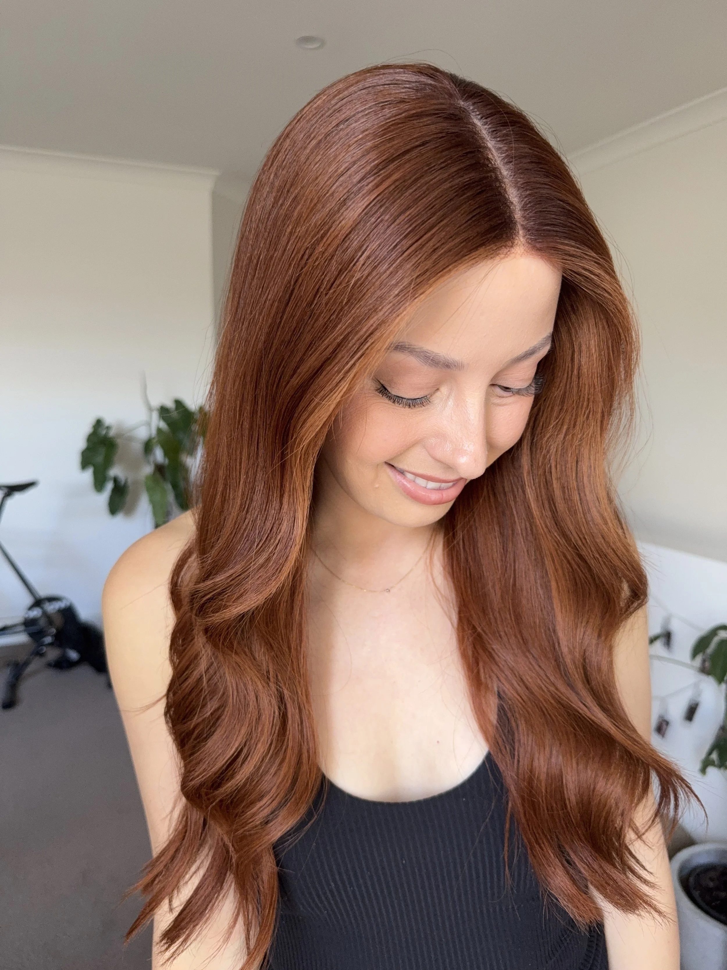 A woman with long, wavy auburn hair smiling downwards in a room with a bicycle and plants in the background.