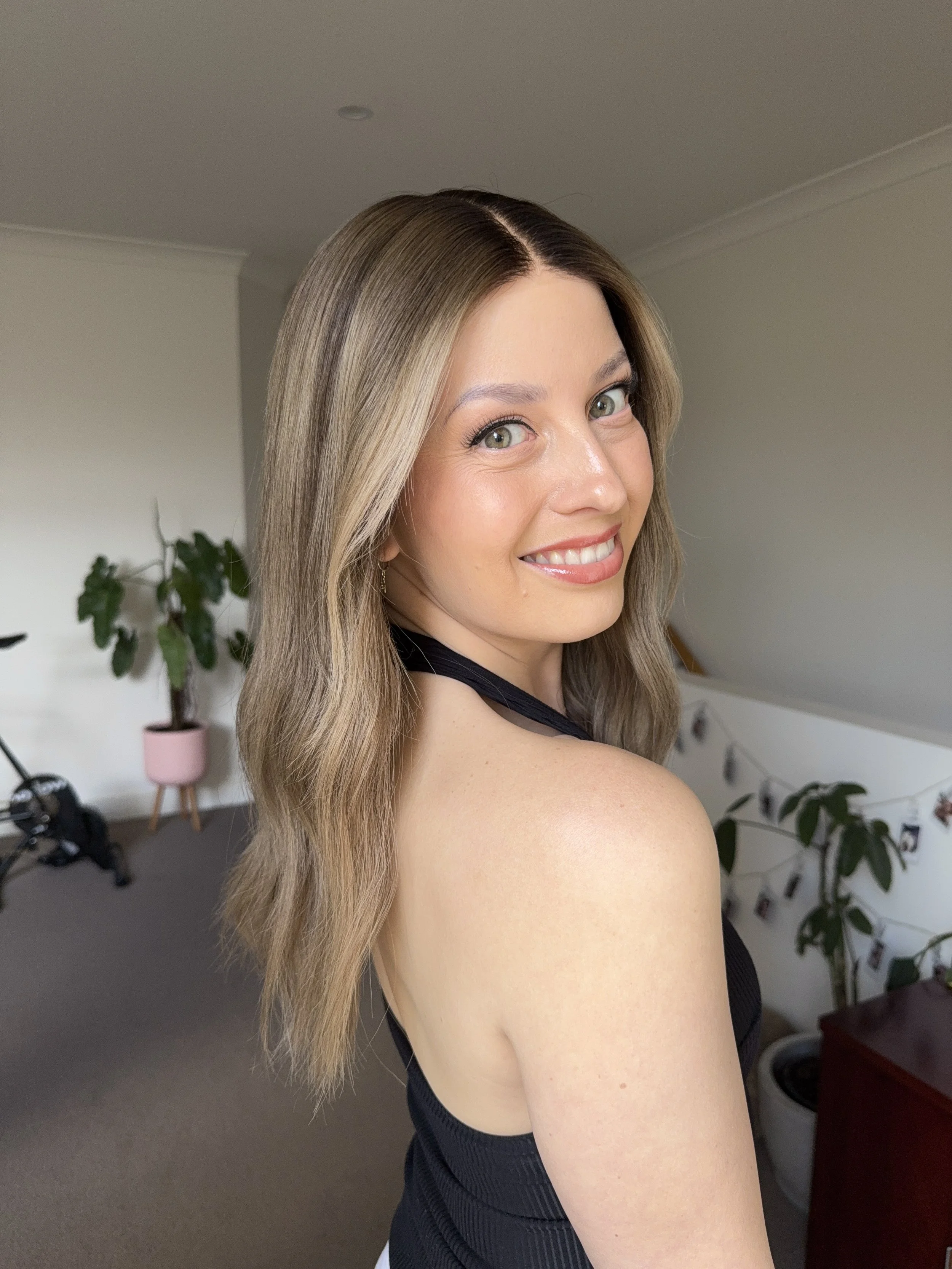 A woman with long, wavy blonde hair smiling at the camera in a room decorated with plants and hanging photos.