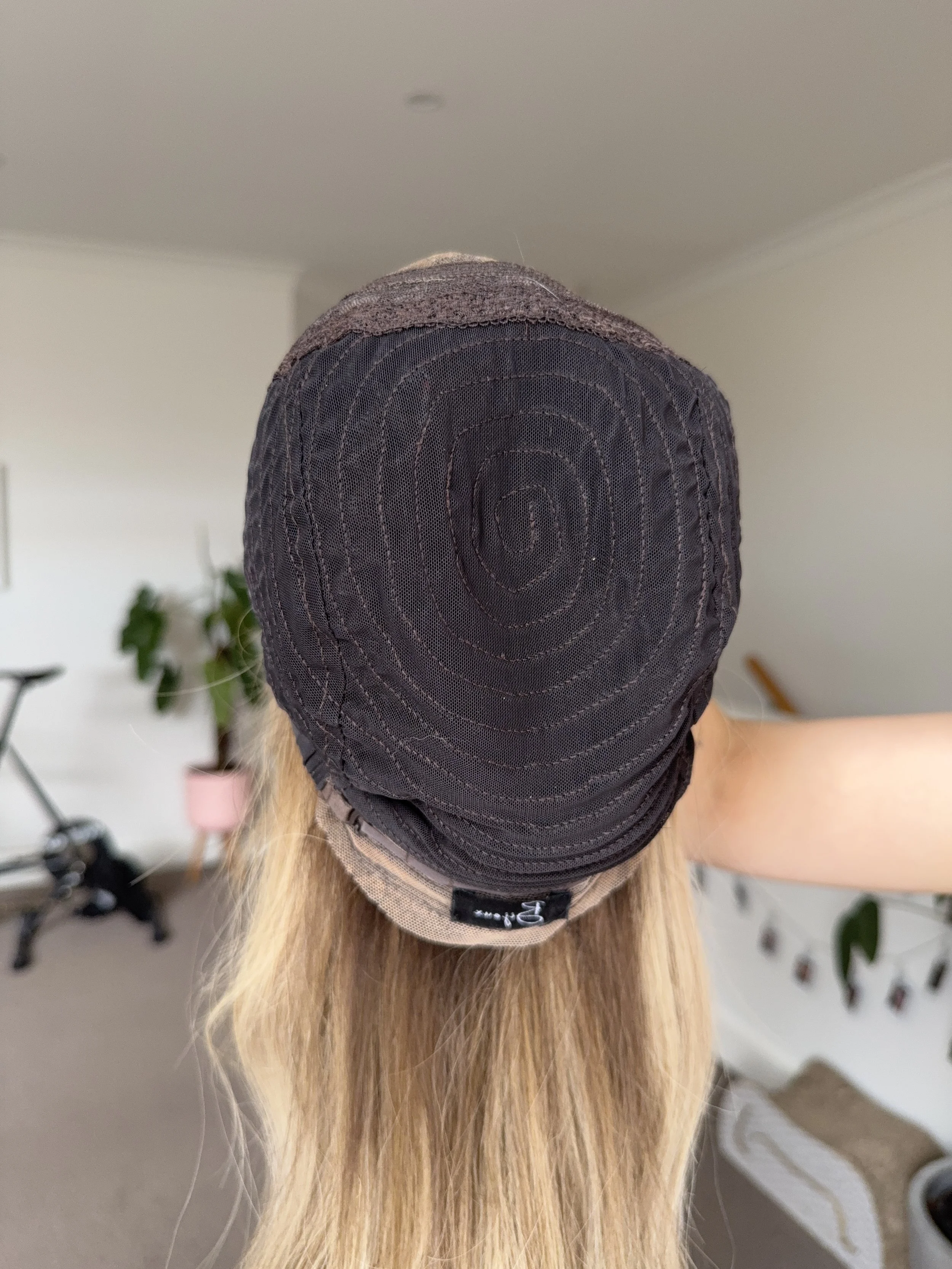 Back of a person's head with long blonde hair, wearing a black hairpiece cap with visible stitching and a small black tag at the edge.