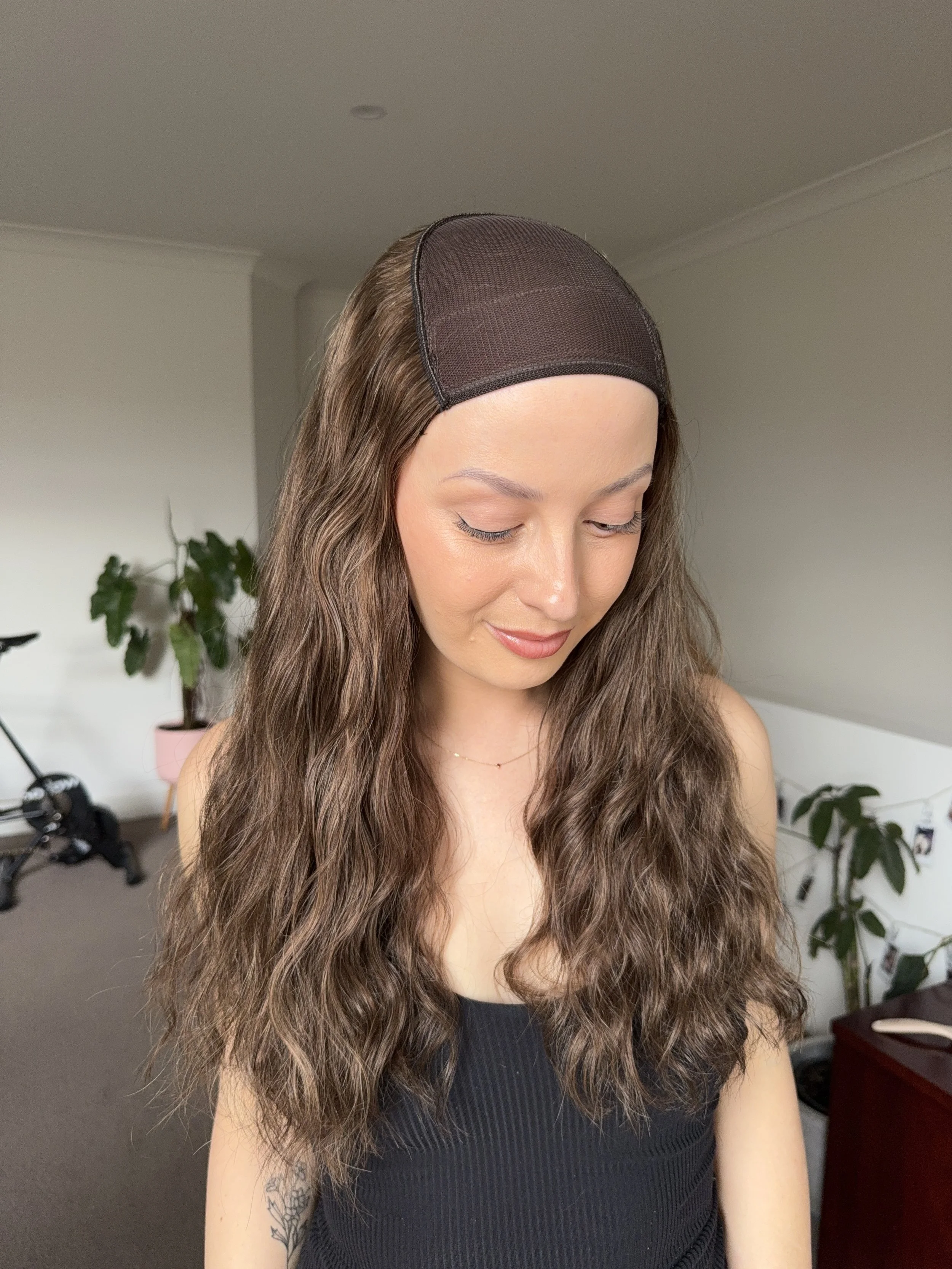 A woman with long wavy brown hair wearing a black wig cap and a black sleeveless top, looking down with a slight smile in a room with plants and furniture.