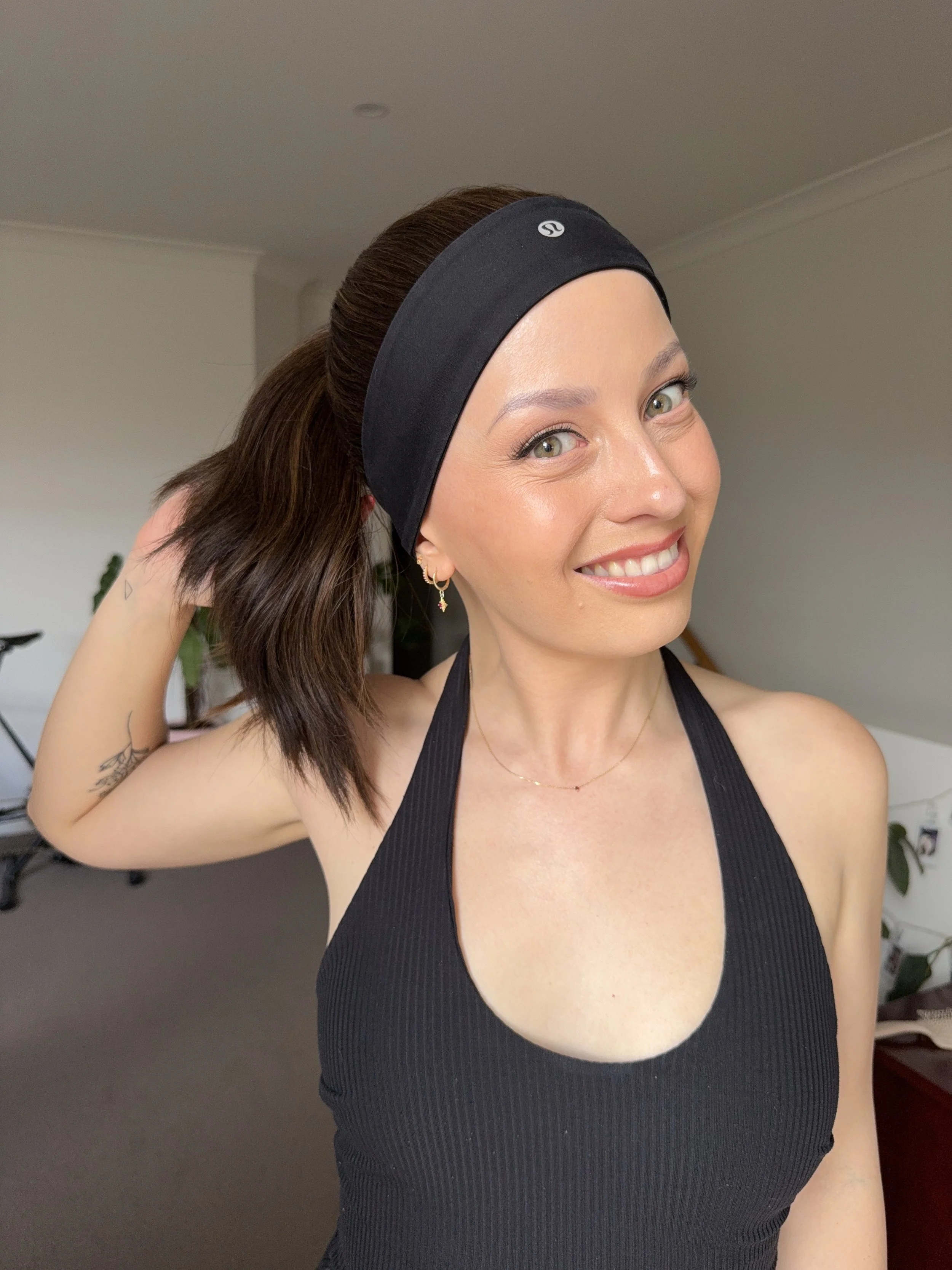 A woman with light skin and brown hair pulled back, wearing a black headband and black athletic top, smiling and holding her hair back with her right hand.