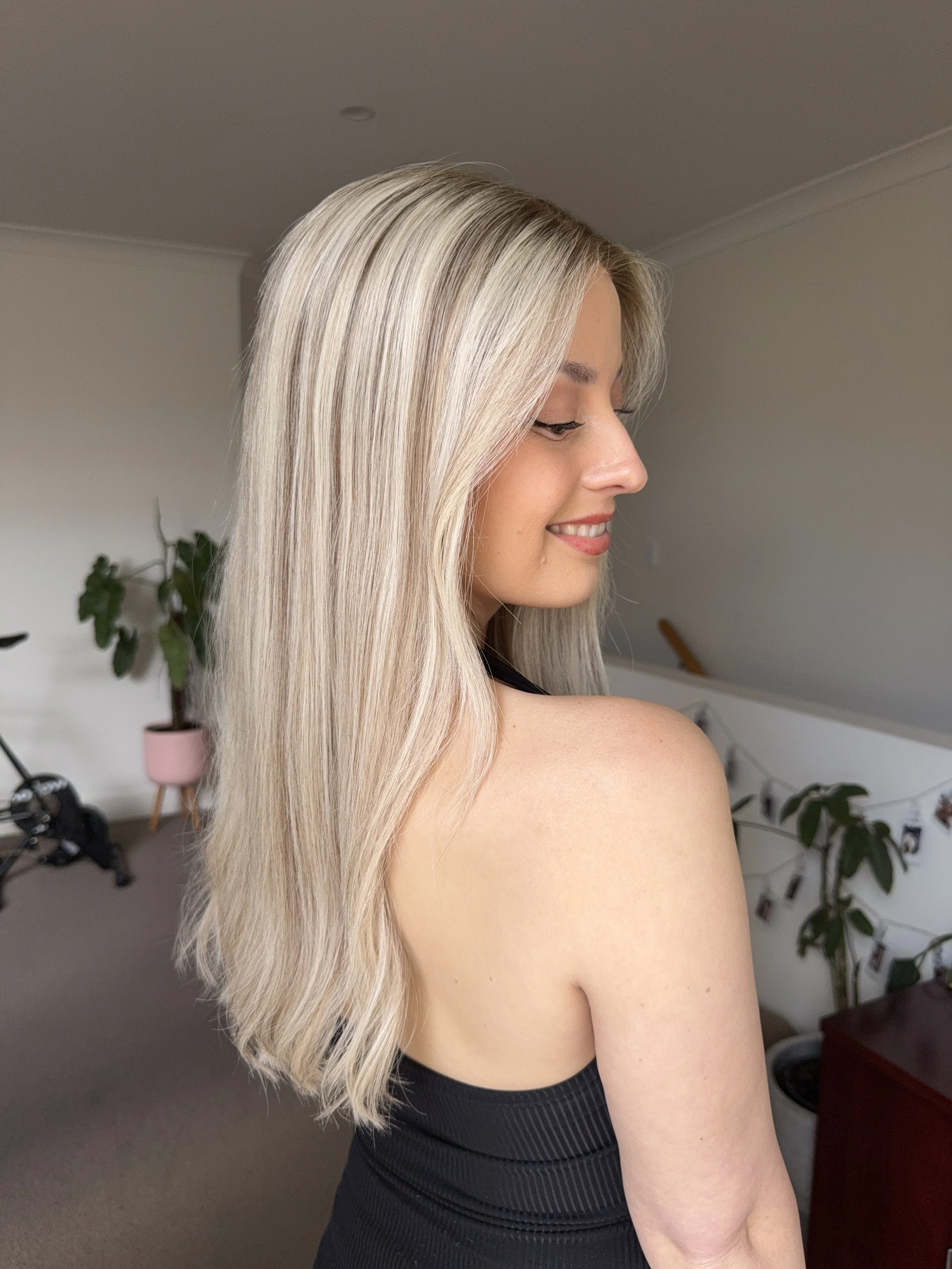 A woman with long, blonde hair smiling and looking down, wearing a black sleeveless top, in a room with plants, a bicycle, and photos hanging on a string in the background.