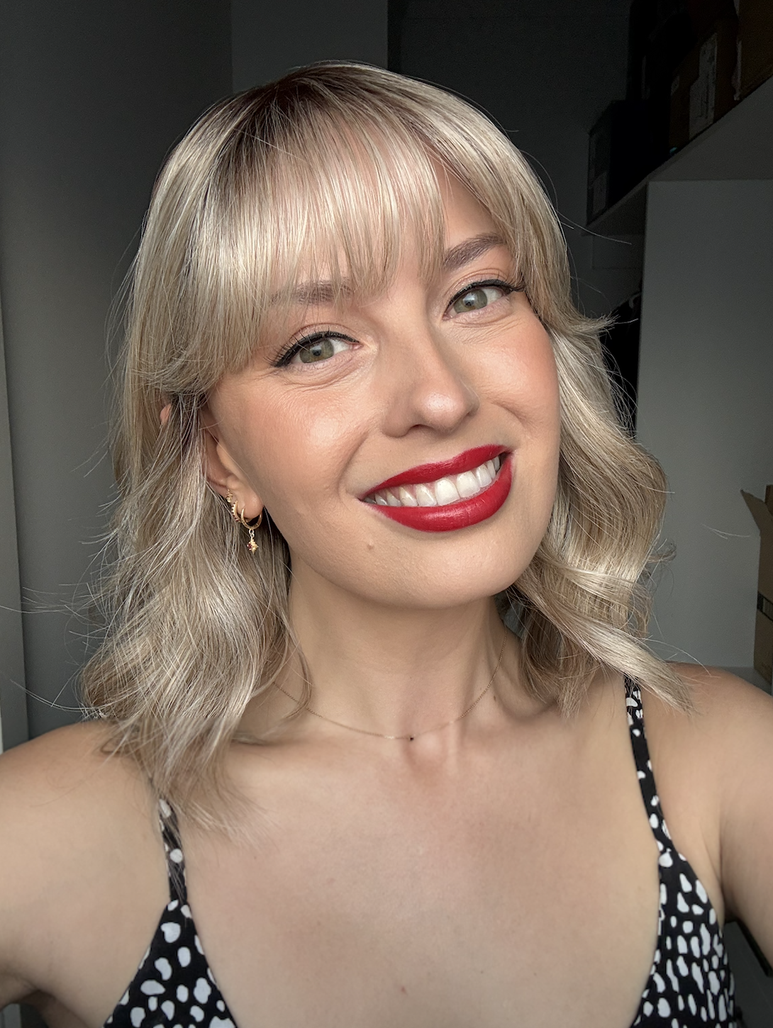 A smiling woman with blonde, wavy hair, red lipstick, and wearing a black and white polka dot top, taking a selfie indoors.