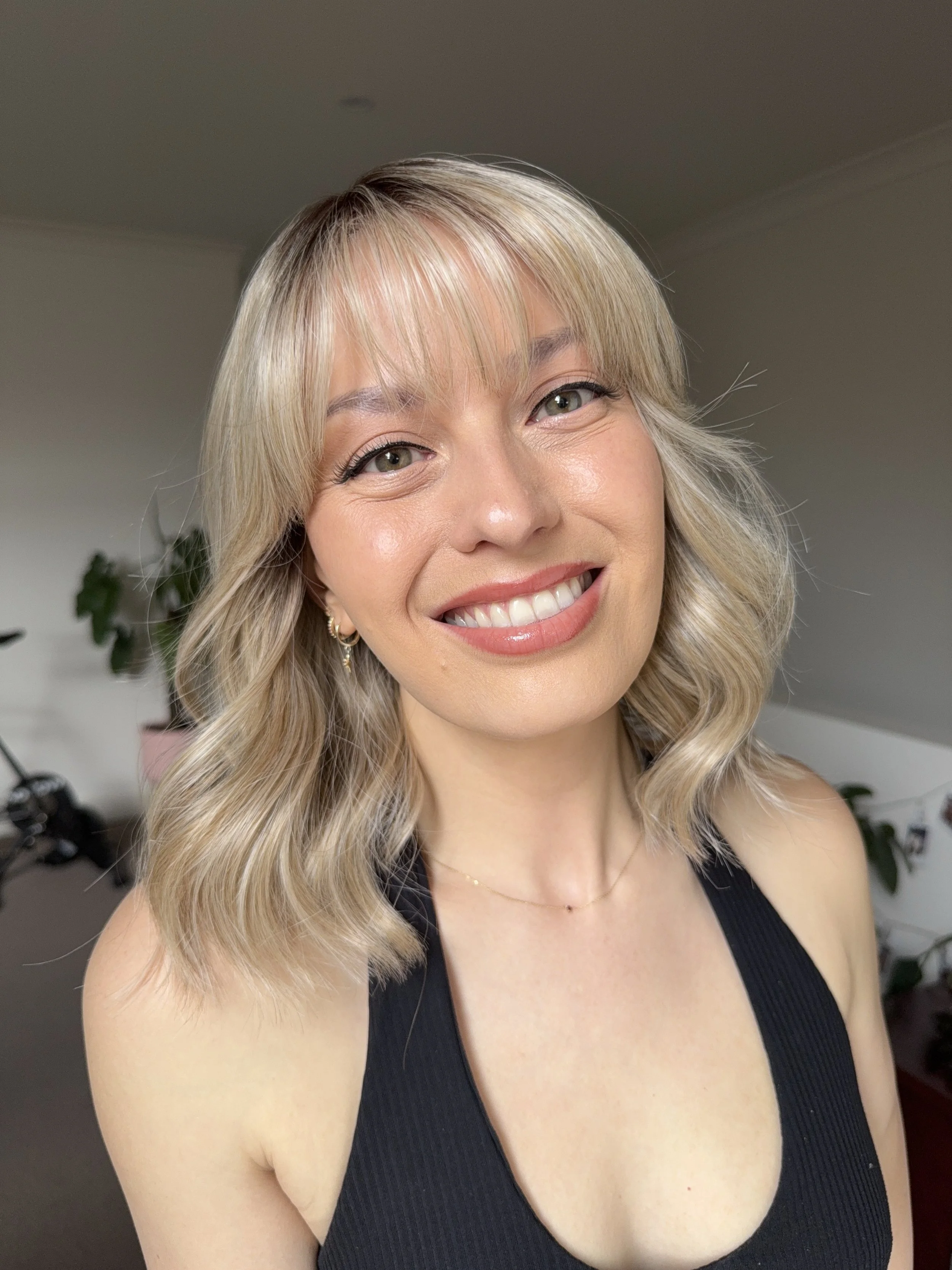 A woman with blonde wavy hair, smiling and wearing a black sleeveless top, standing indoors with plants and furniture in the background.