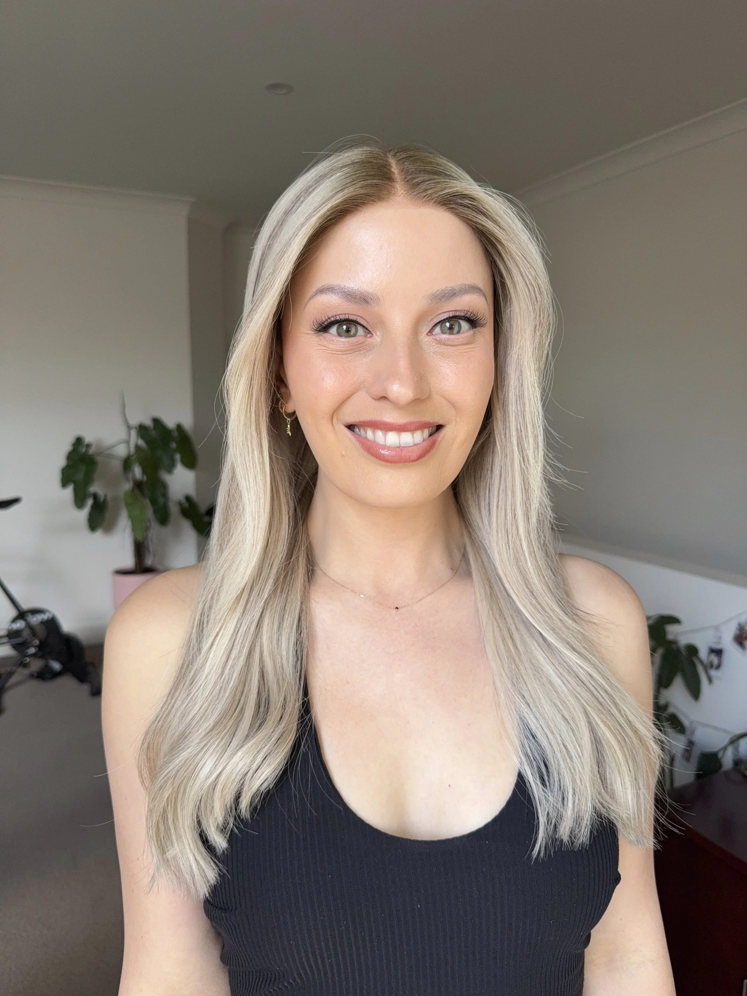 A young woman with long blond hair, wearing a black sleeveless top and a delicate necklace, smiling in a well-lit room with plants in the background.
